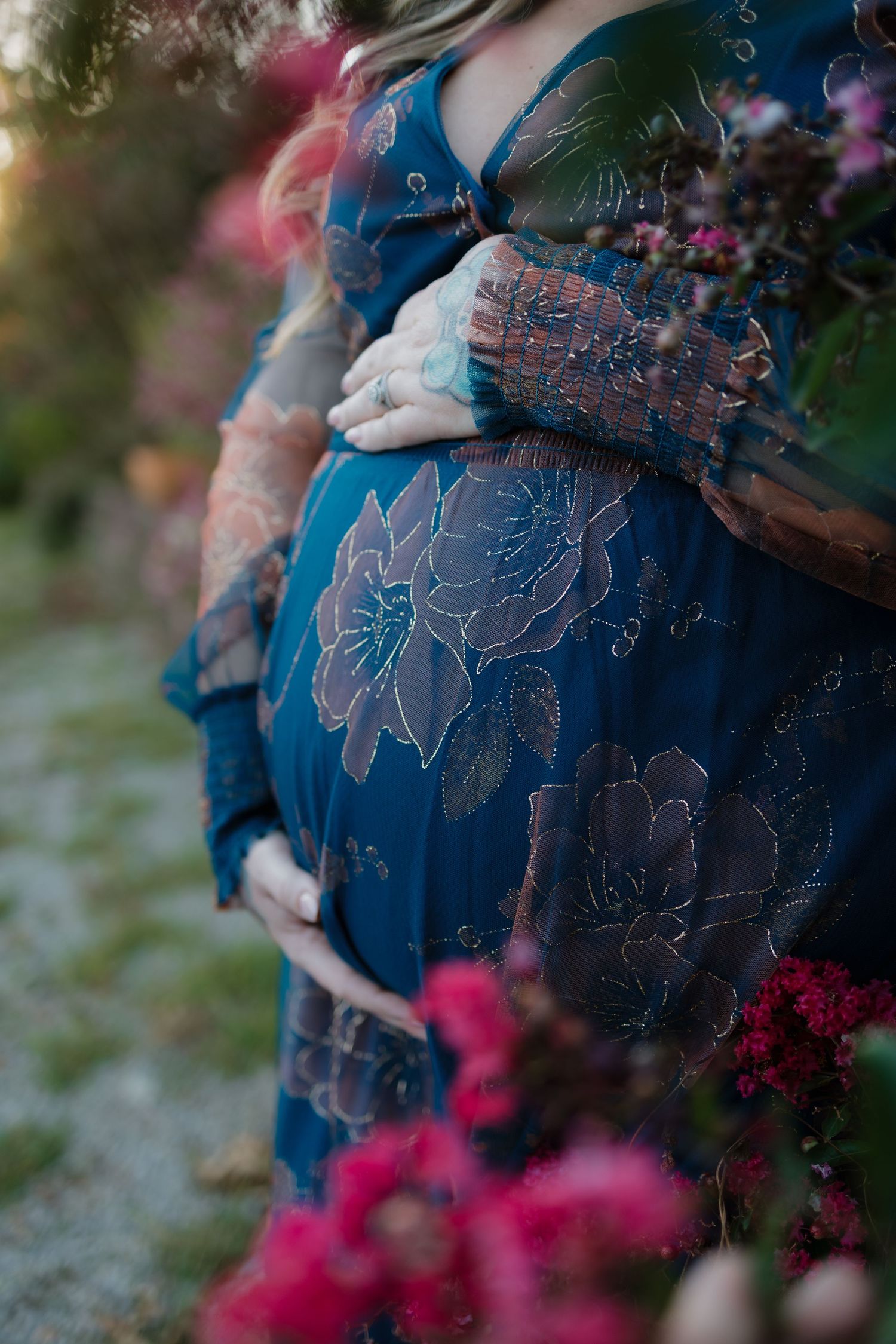 Pregnant woman in teal floral dress cradling baby bump outdoors at golden hour surrounded by pink flowers.
