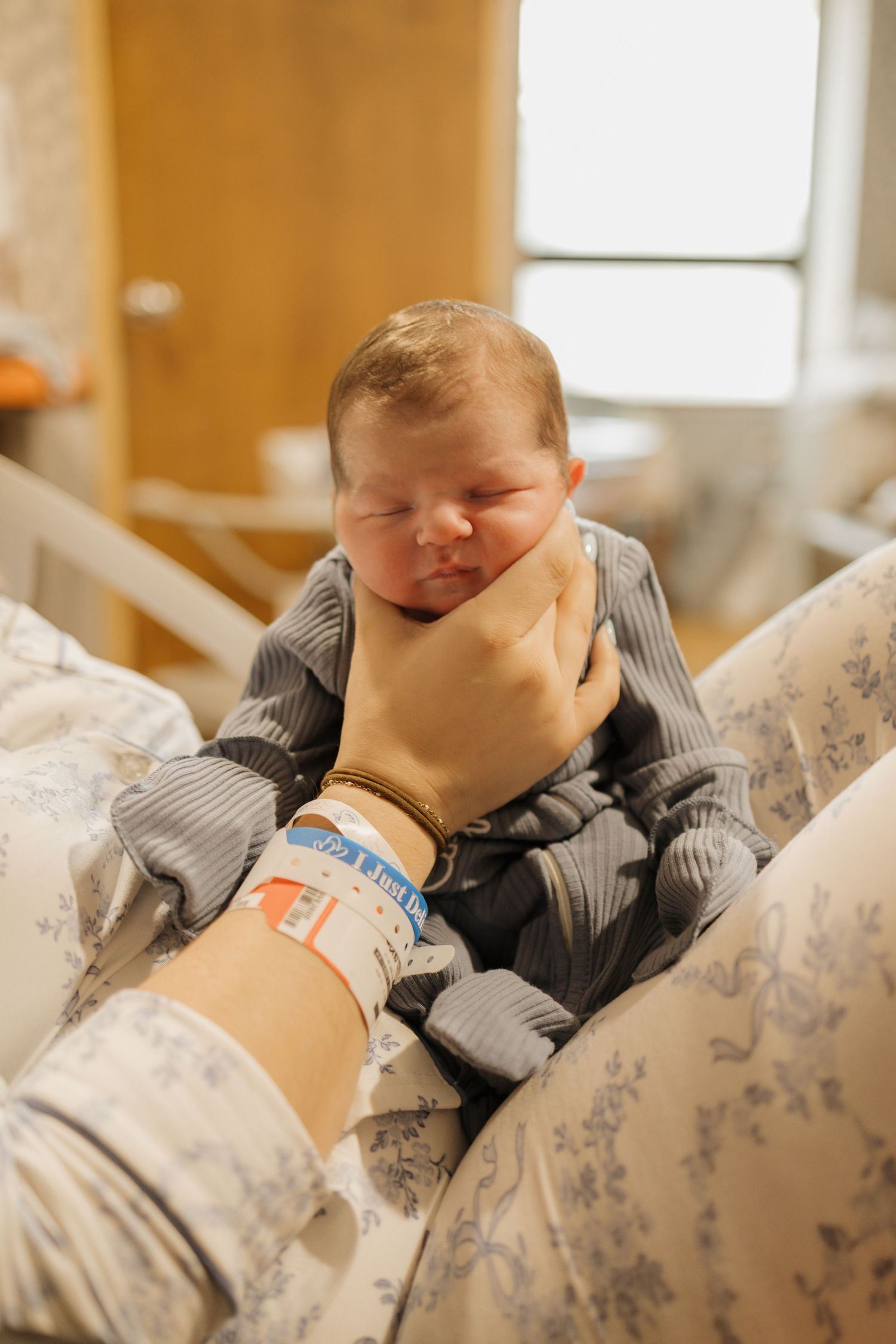 A newborn baby in a gray sleeper being cradled by a hospital-banded parent's hand in a maternity room.