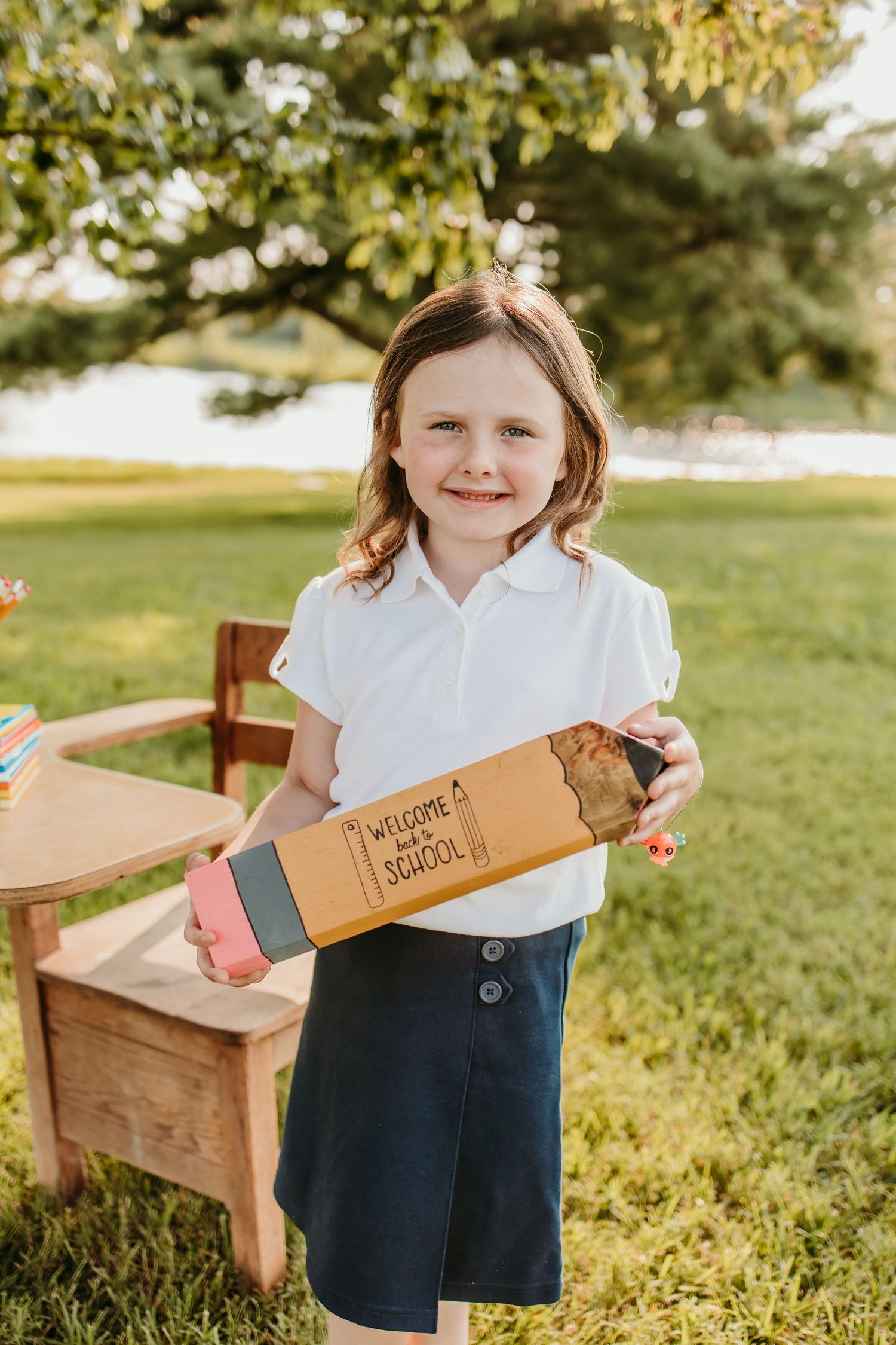 Young girl in white polo smiling outdoors, holding a Welcome Back to School pencil-shaped gift box.