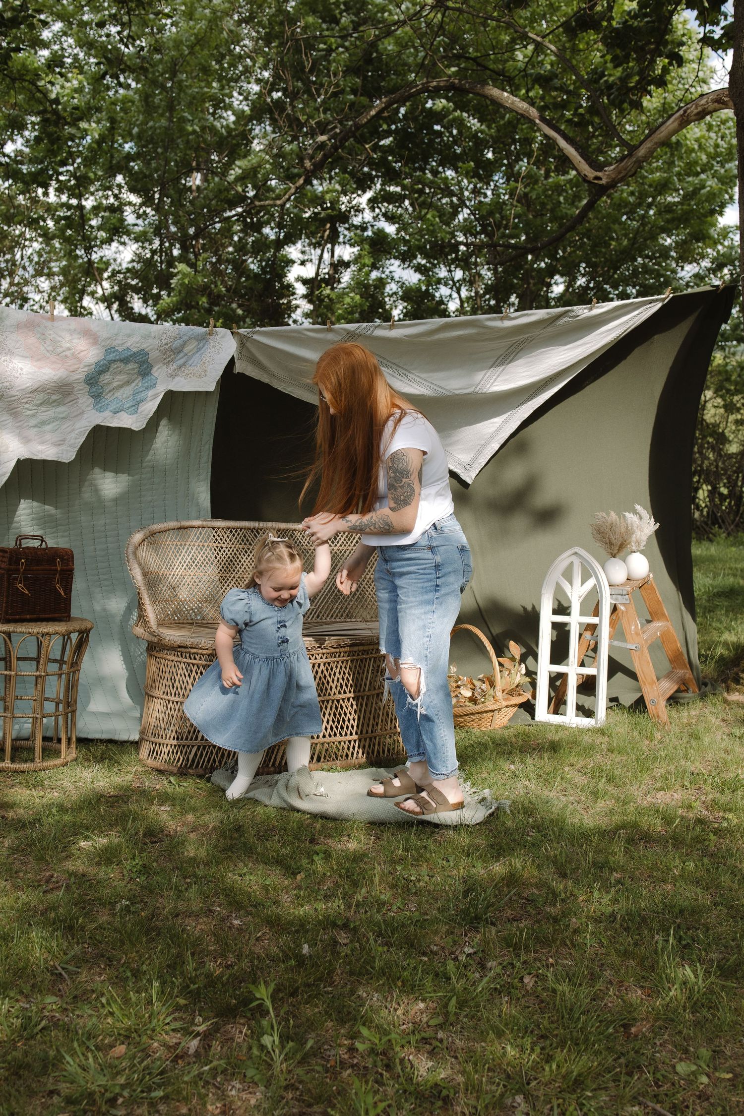 Red-haired tattooed mother in jeans helps toddler girl in blue dress near wicker chair outdoors.