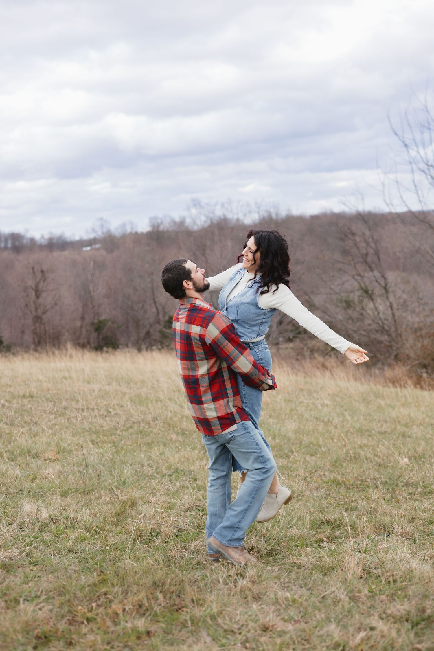 Man joyfully lifting woman in denim overalls in open grassy field under cloudy sky.