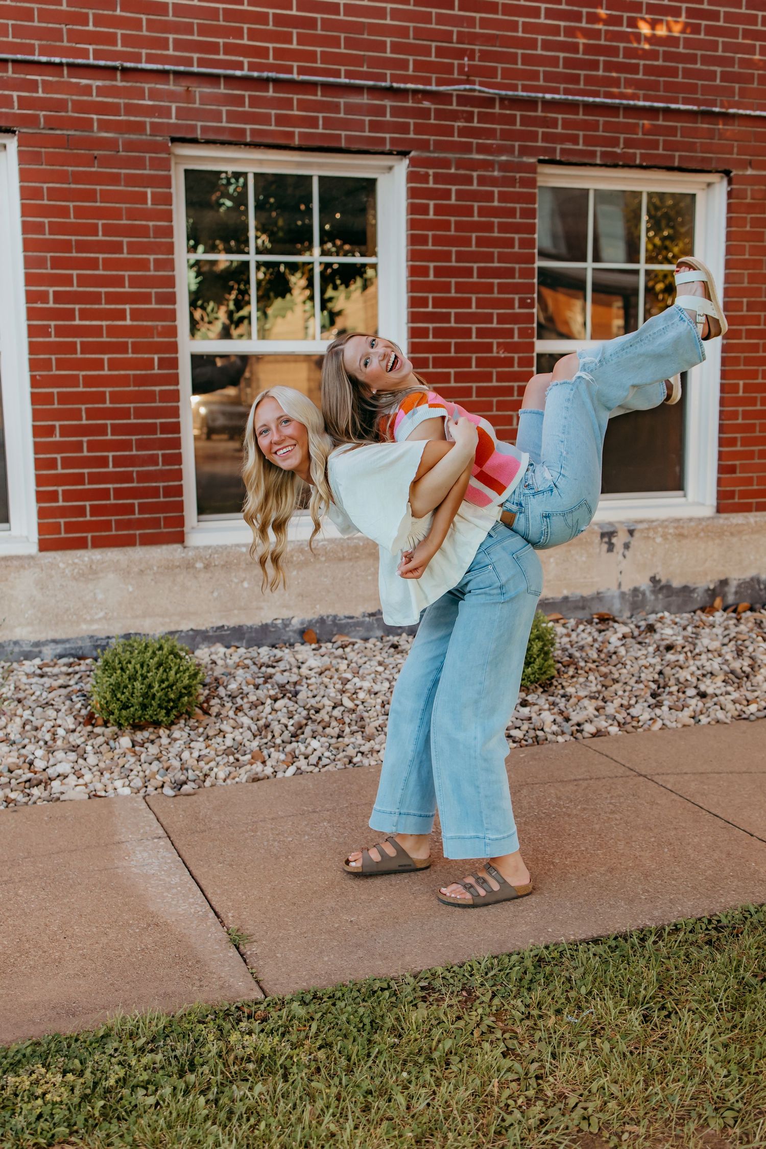 Two seniors laughing outdoors, one giving the other a piggyback ride in front of a red brick building.