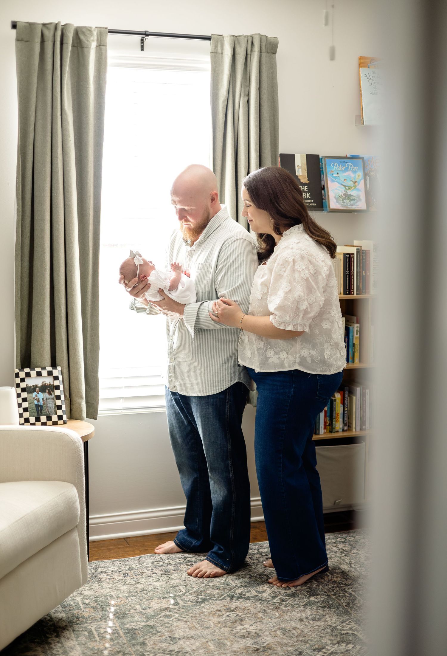 A couple stands by a bright window holding their newborn baby in a cozy room with bookshelves.
