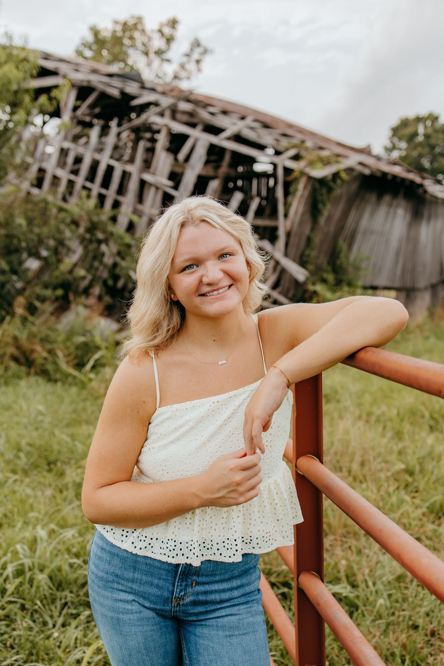 Young blonde woman in white top and jeans leaning on red farm gate with rustic barn in background.