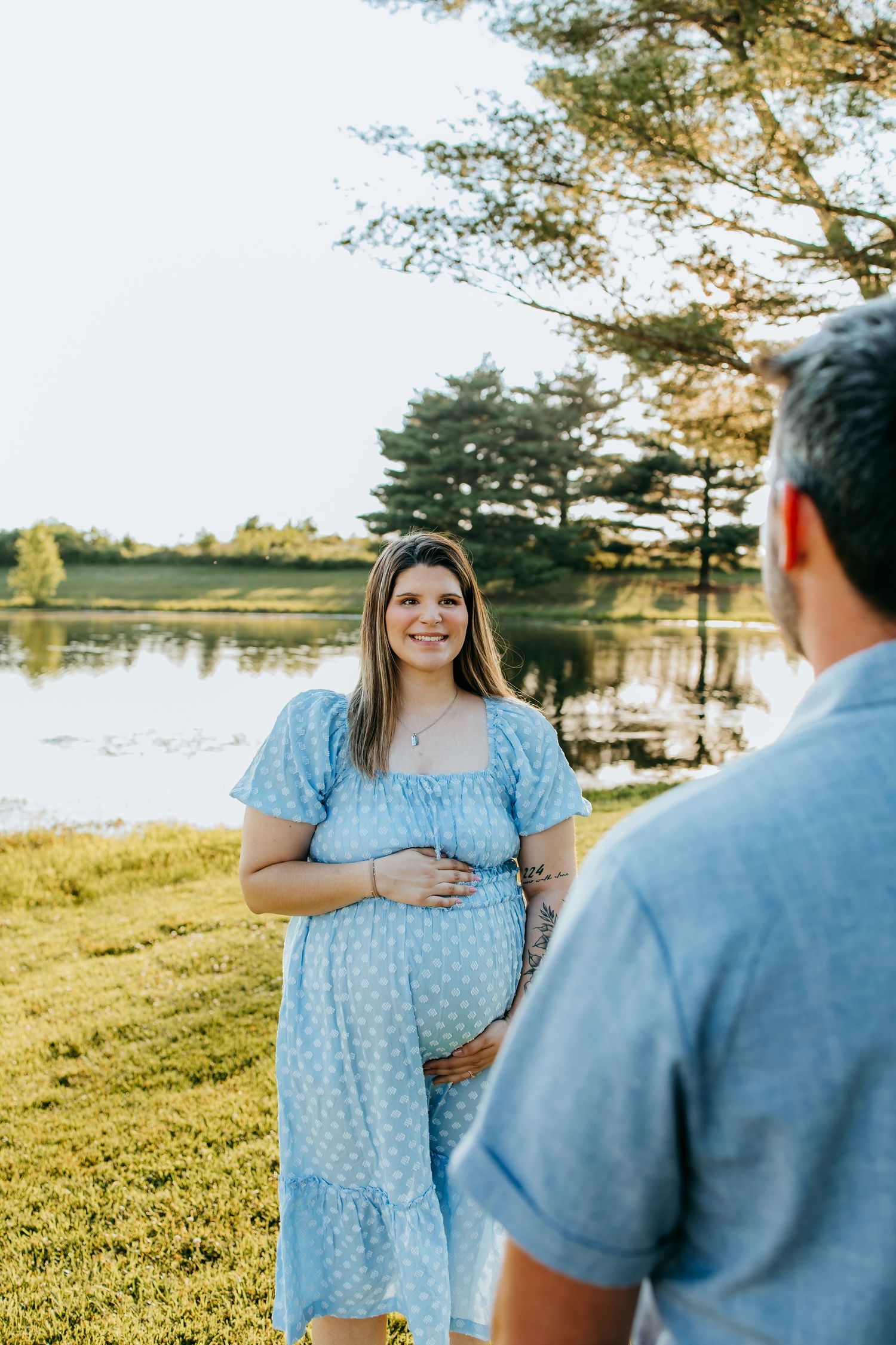 Smiling pregnant woman in blue floral dress holds baby bump near a serene lake during golden hour maternity photoshoot.
