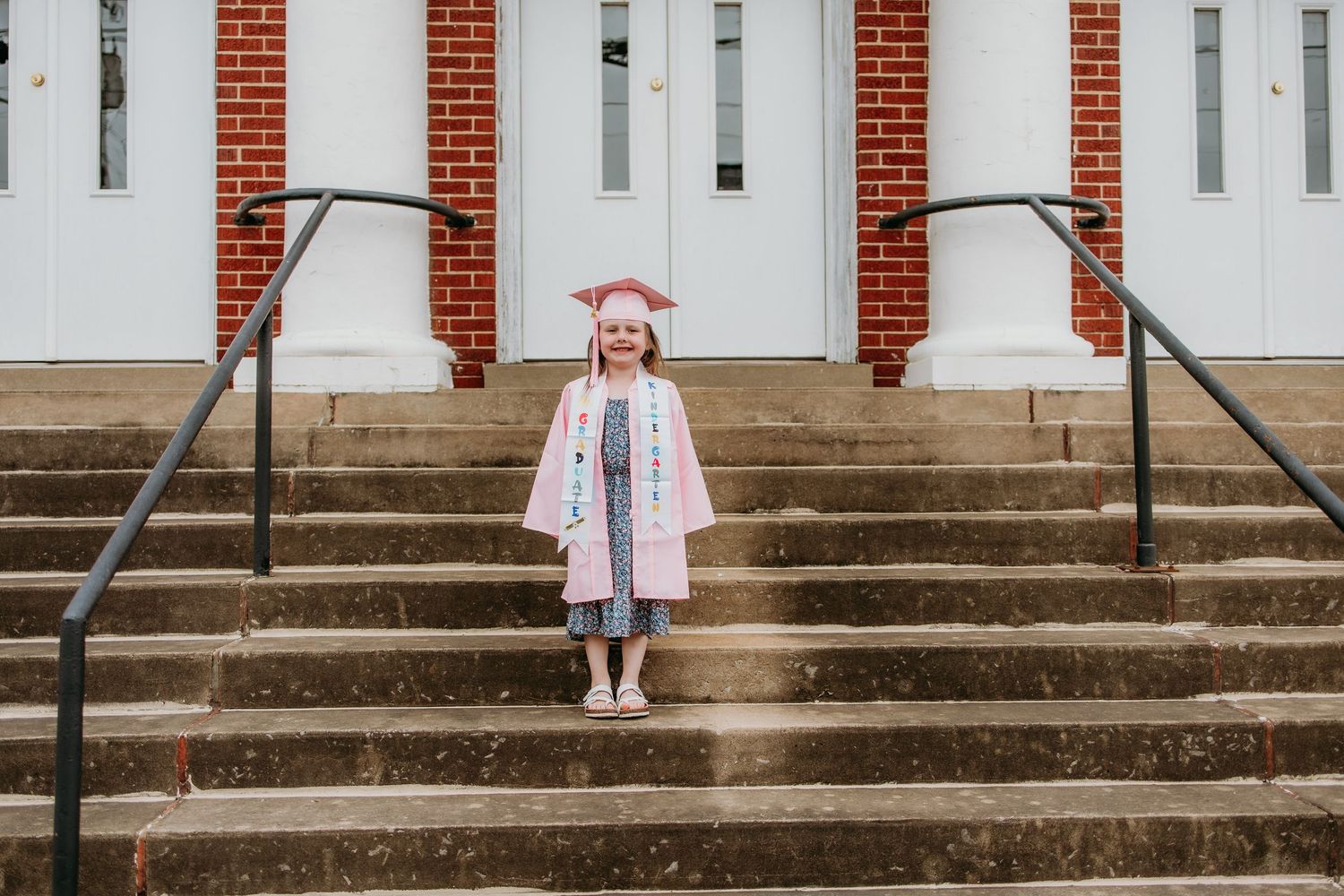 Young girl in pink graduation cap and gown smiling on brick steps outside a white building.