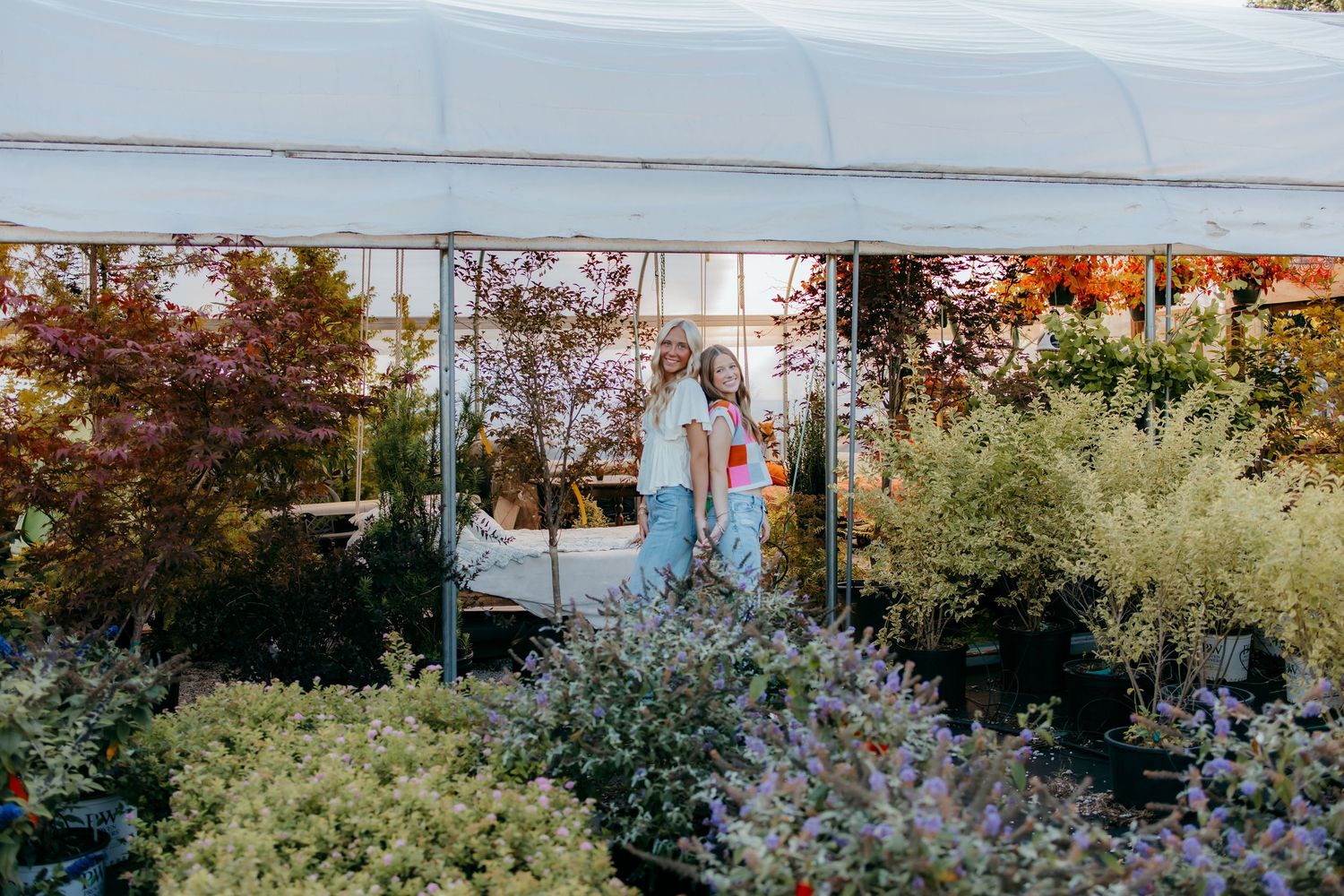 Two women browse plants at an outdoor garden nursery surrounded by lush greenery and colorful shrubs under a white tent.