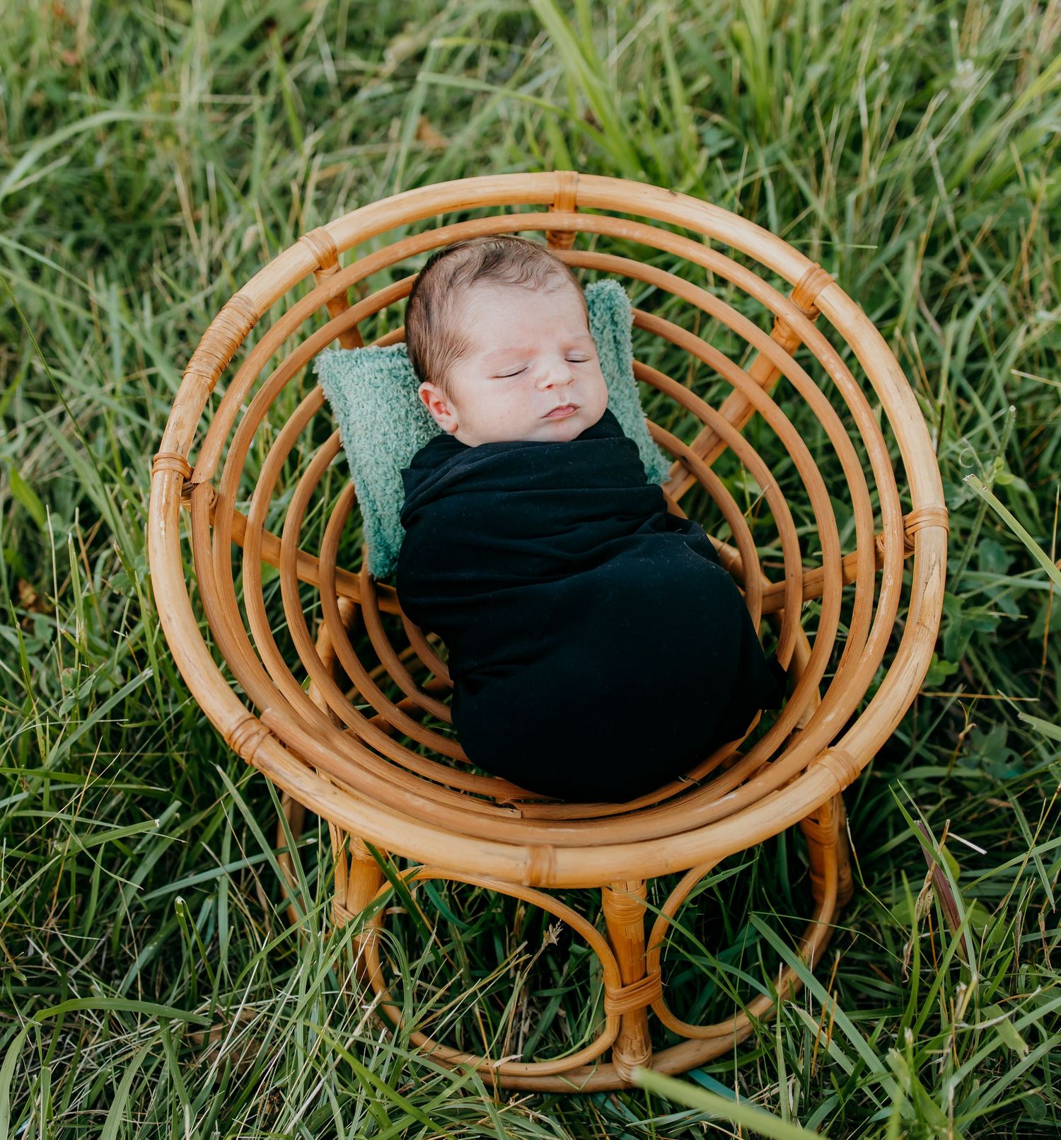 Newborn baby swaddled in black wrap sleeping in a rattan chair surrounded by green grass outdoors.