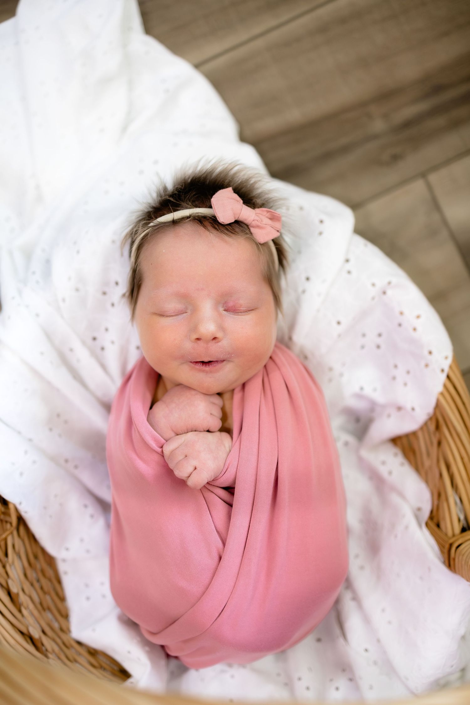 Newborn baby girl swaddled in pink wrap with floral headband sleeping peacefully in a wicker basket.