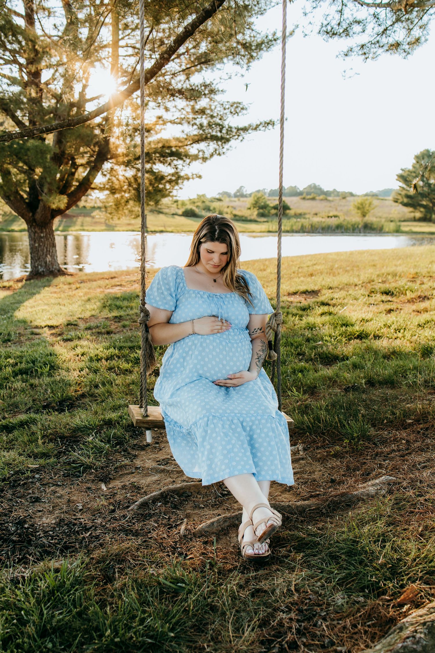 Pregnant woman in blue polka dot dress sitting on wooden swing near lake at golden hour sunset.