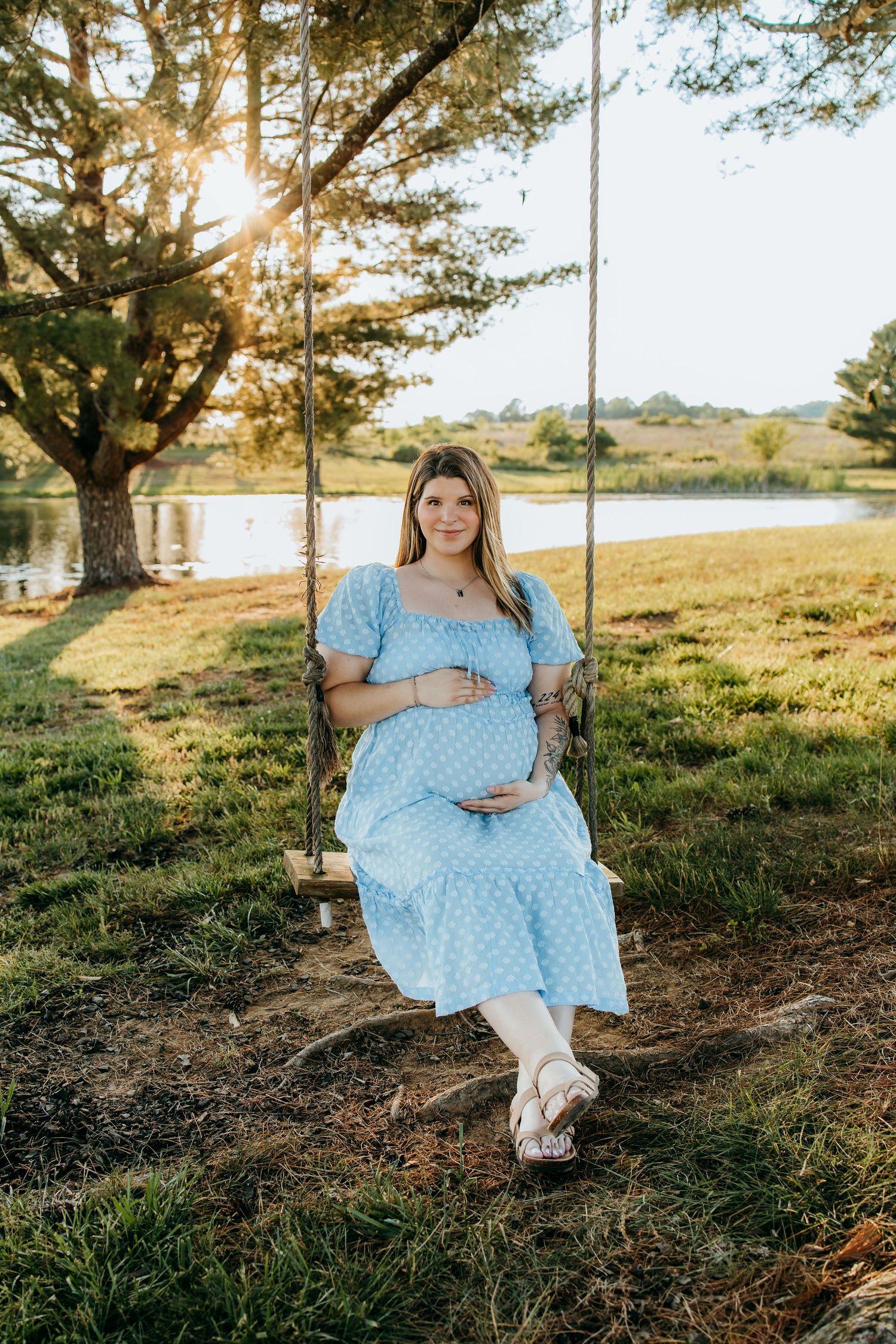 Pregnant woman in blue floral dress sitting on wooden swing near pond at golden hour sunset.