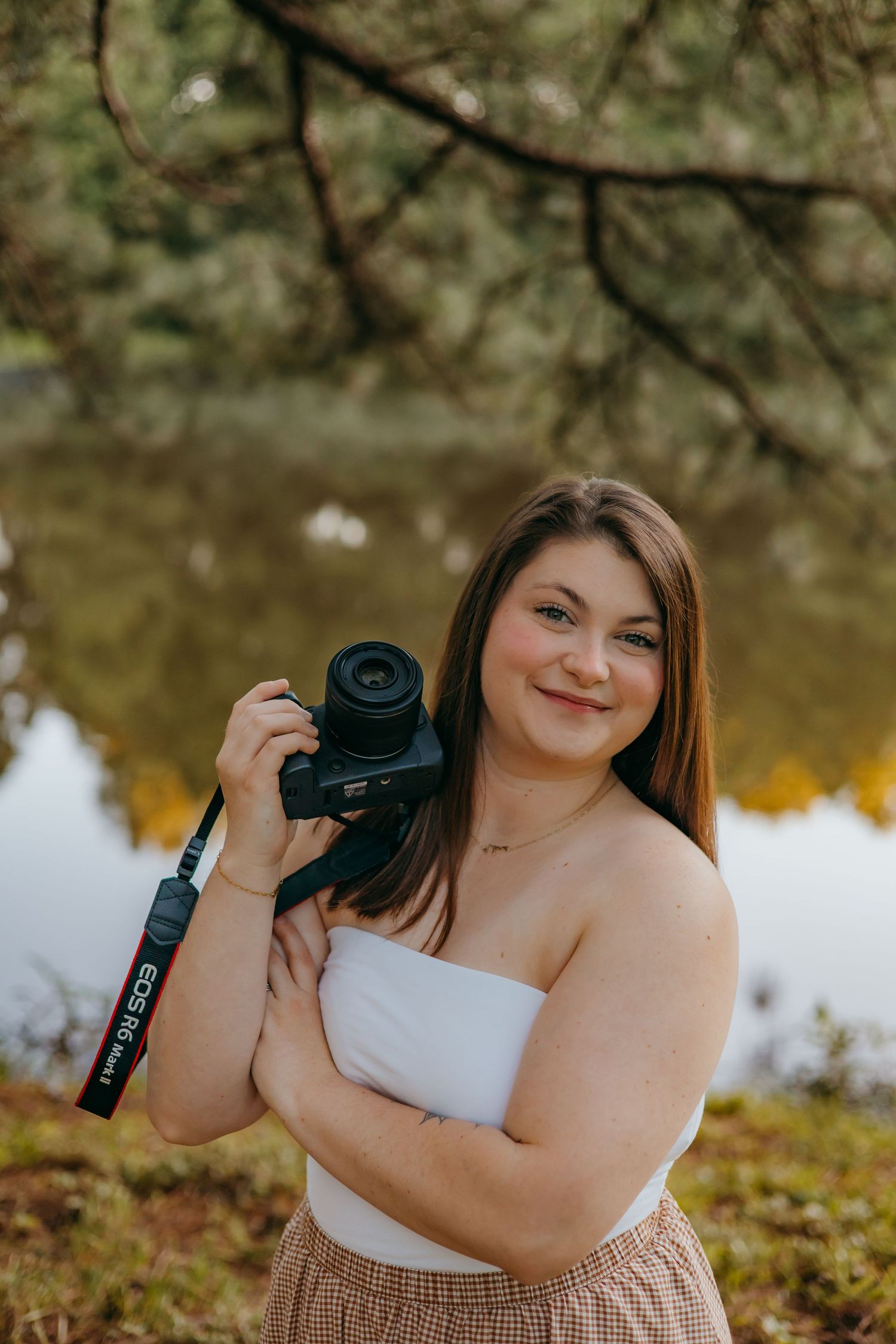 Young woman smiling outdoors holding a Canon DSLR camera near a reflective pond surrounded by autumn foliage.