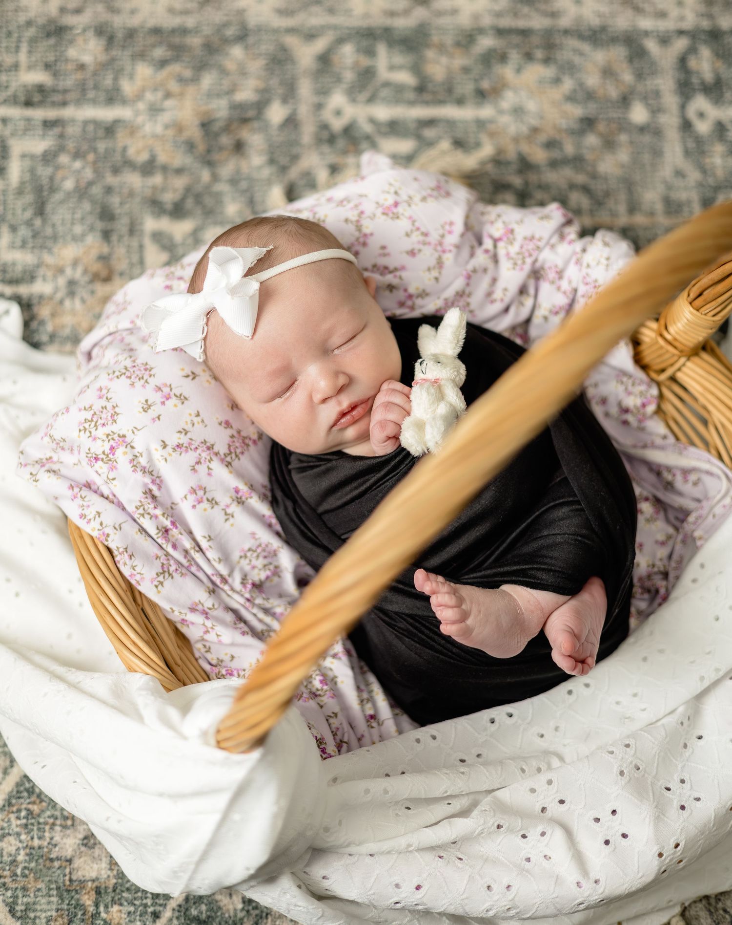 Sleeping newborn baby girl in a wicker basket, wrapped in black, wearing a white bow headband, holding a bunny toy.