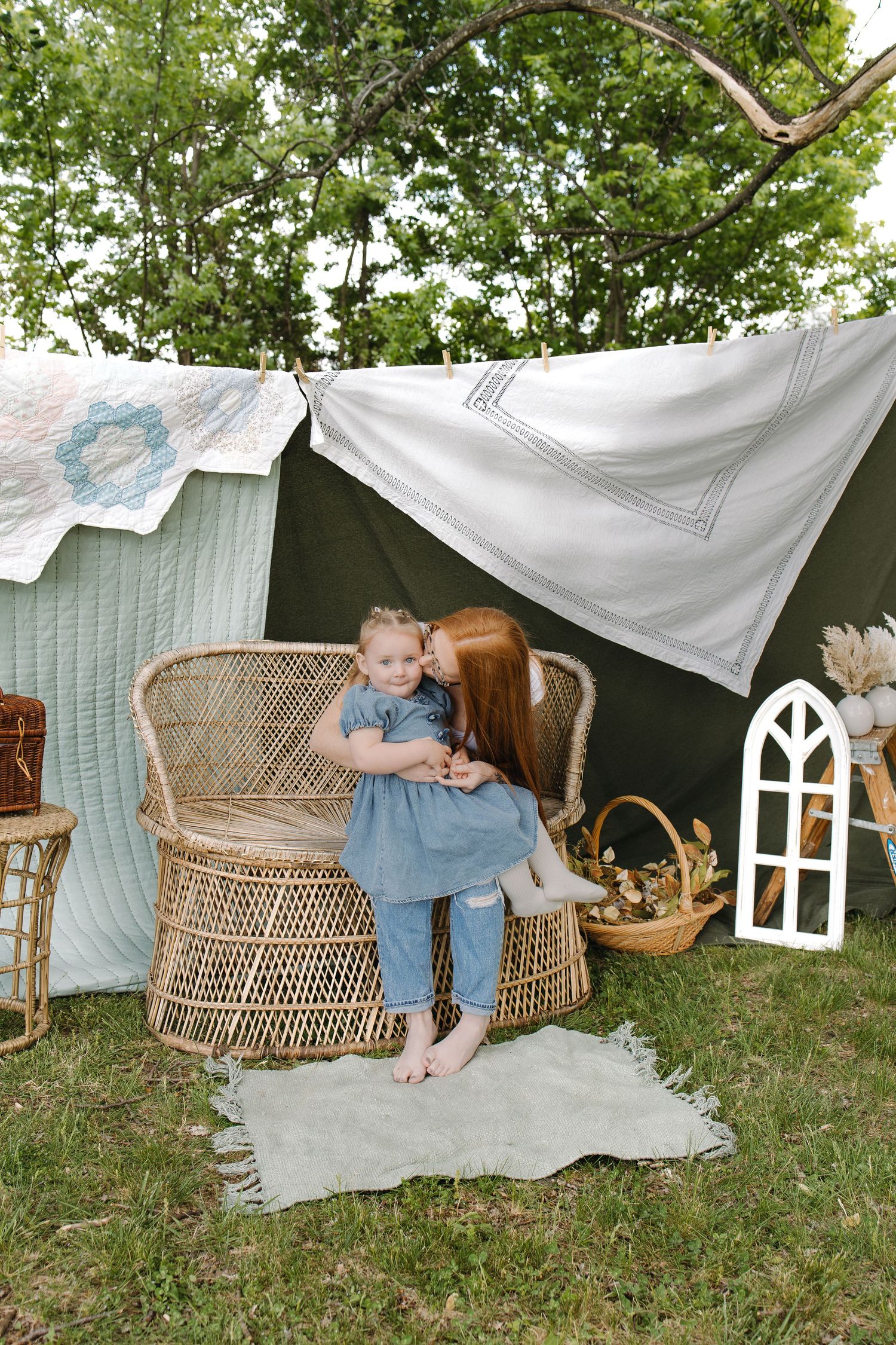 A redheaded woman kisses a young girl in a blue dress beside wicker chairs under a cozy outdoor tent setup.