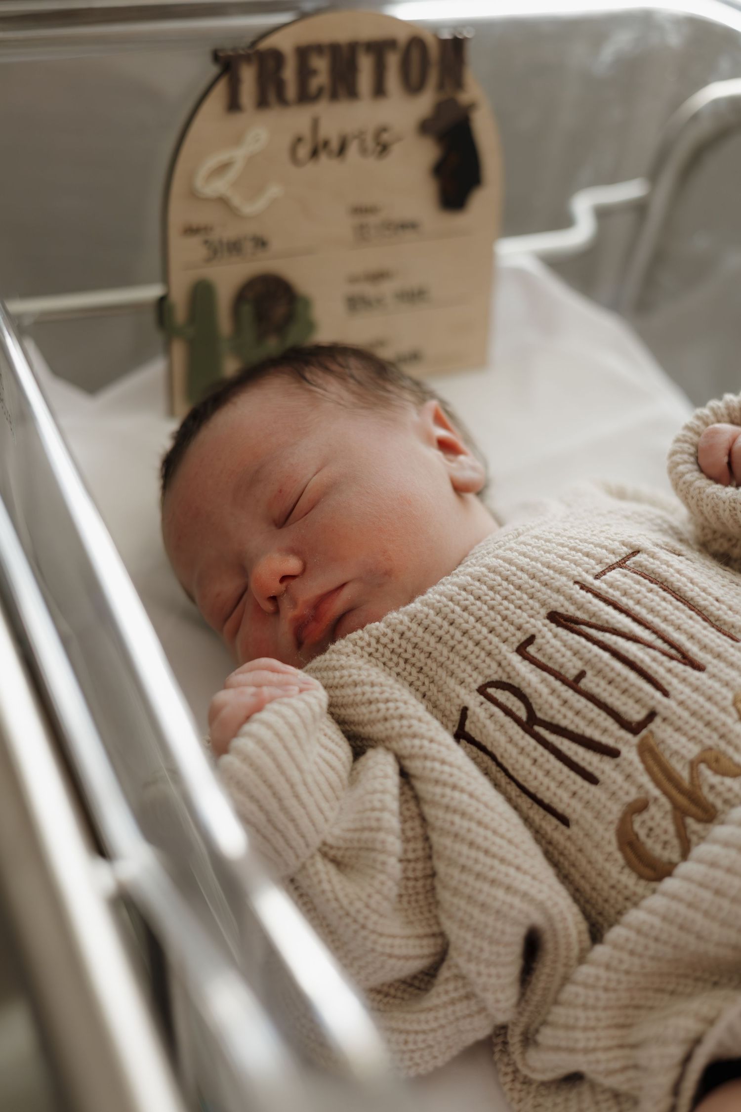 Newborn baby Trenton Chris sleeping peacefully in hospital bassinet wearing personalized knit onesie with name sign.