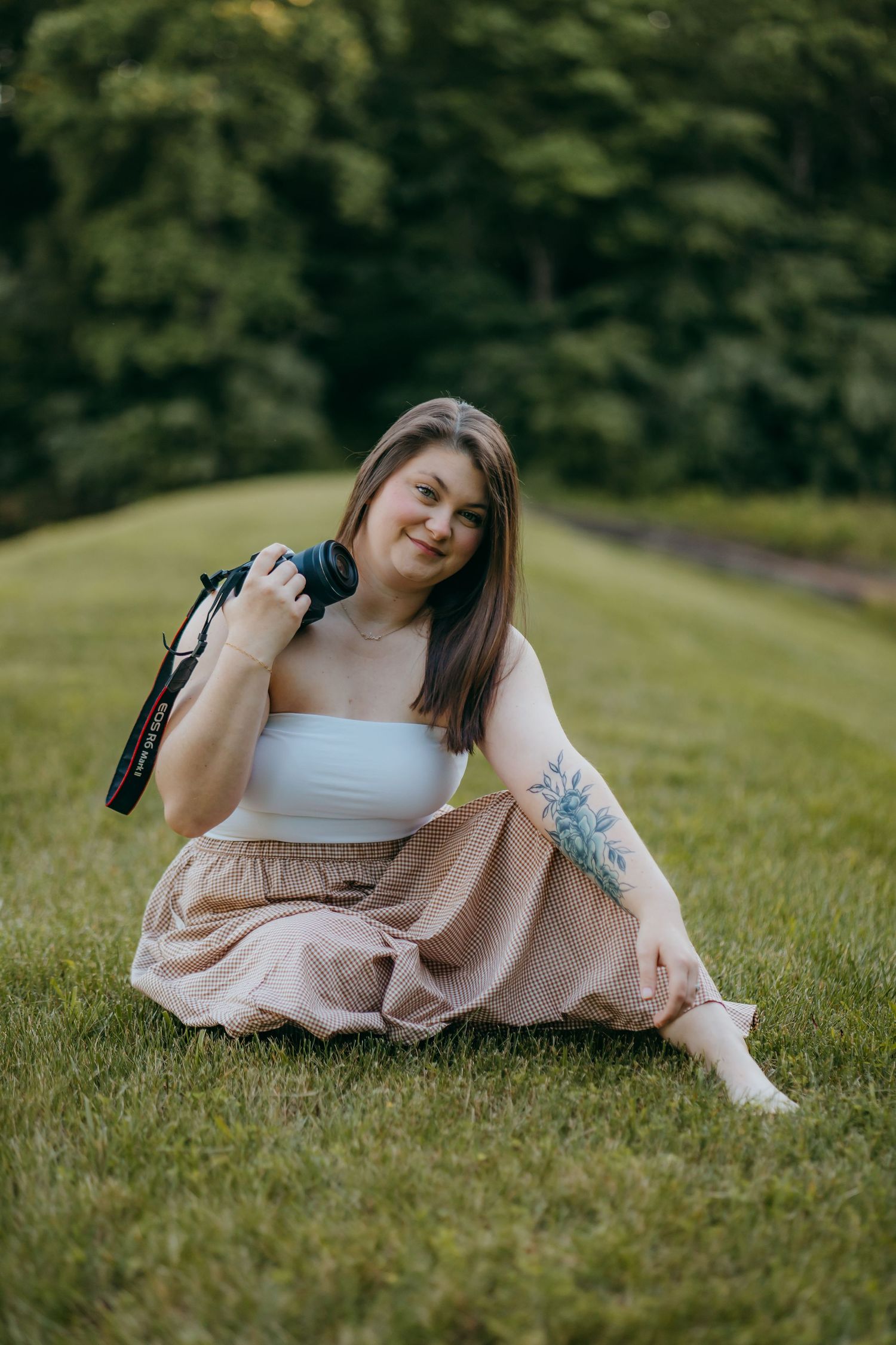 Young woman with tattoo sits on grass holding camera, wearing white tube top and beige skirt in park.