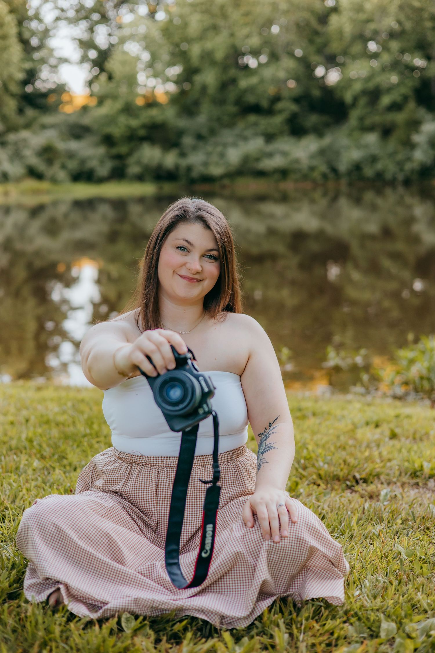 Woman sitting on grass holding Canon camera toward viewer near a serene pond surrounded by lush green trees.