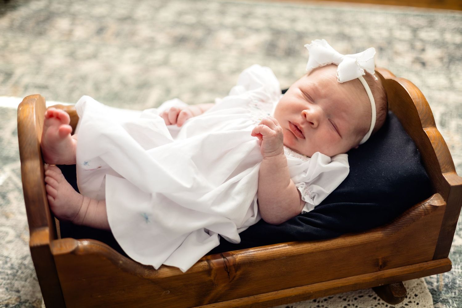 Newborn baby in white dress and bow headband sleeping peacefully in a small wooden bed.