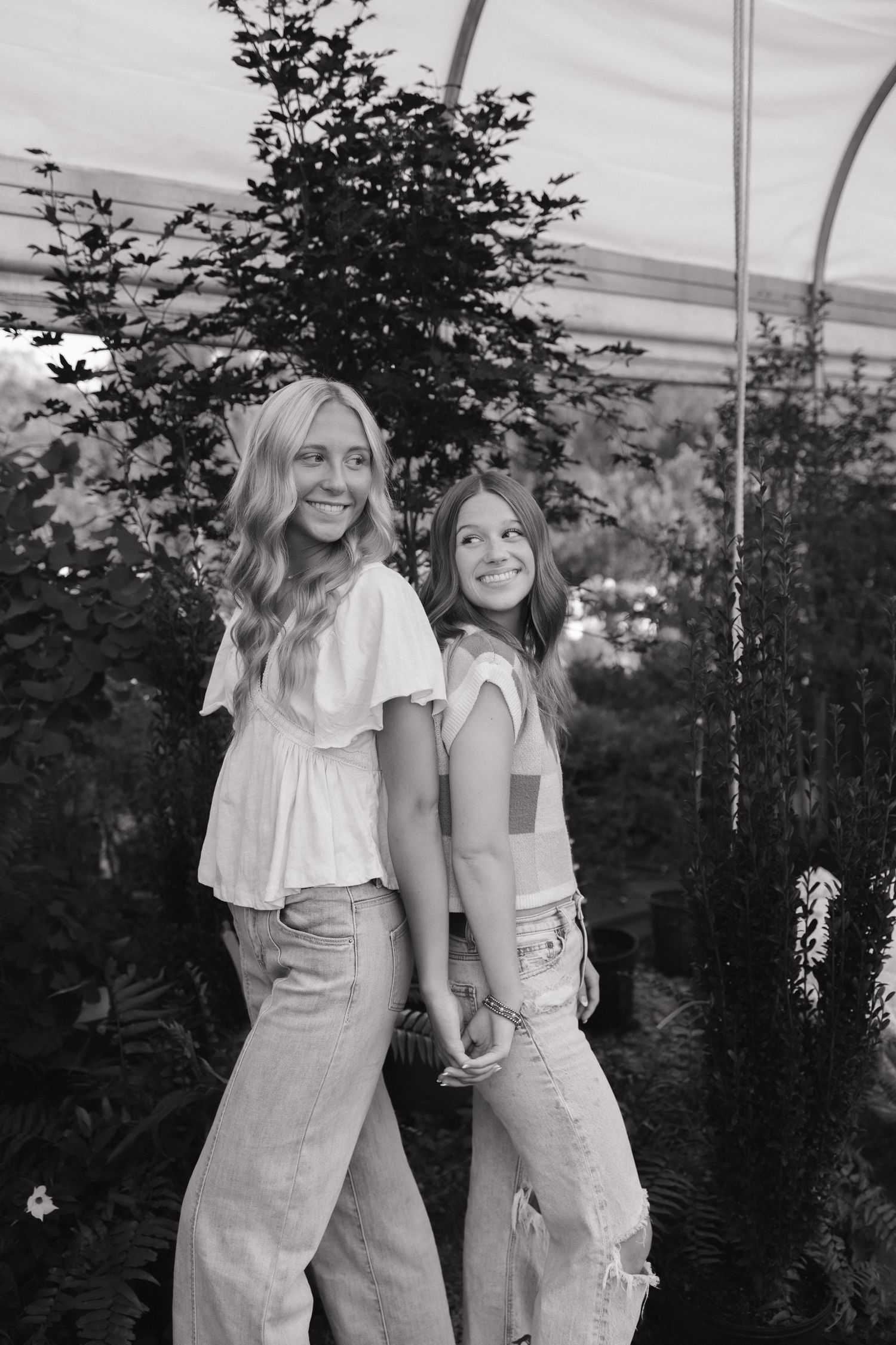 Two seniors smile and hold hands in a greenhouse, wearing jeans and light tops in a black and white photo.