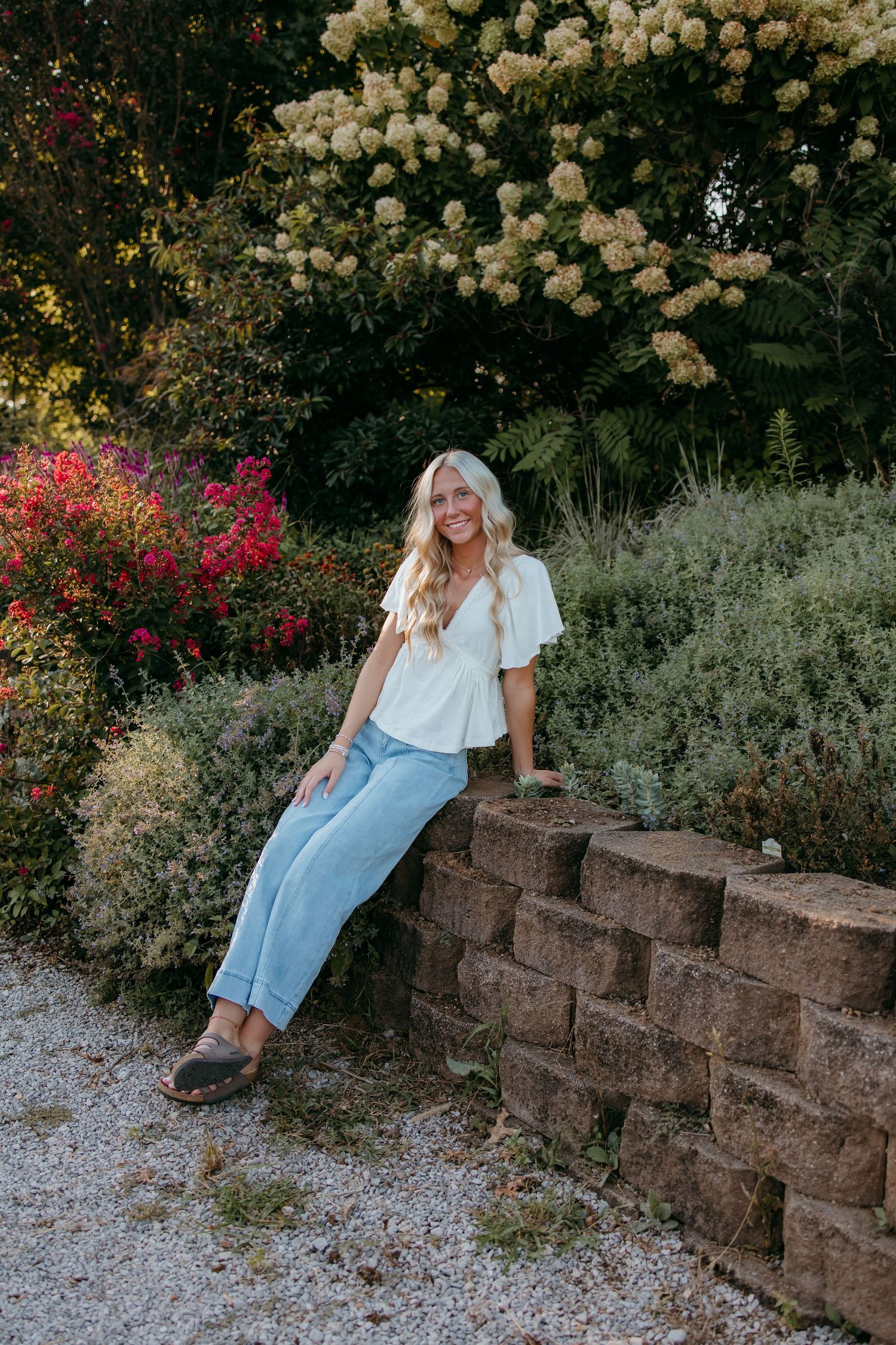Smiling senior in white top and light jeans sitting on stone wall surrounded by colorful garden flowers.