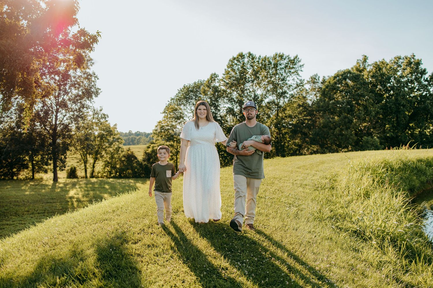 Family of three walking on lush green grass in golden hour sunlight, woman in white dress, man and child in casual attire.