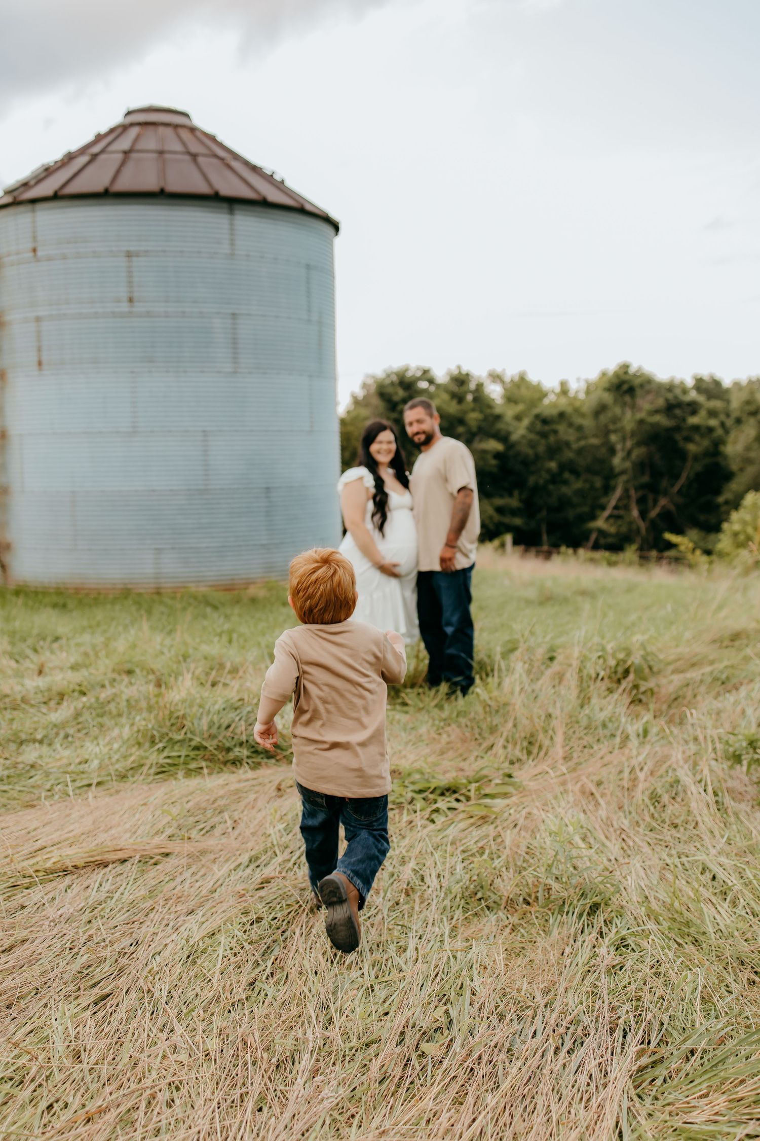 A young red-haired boy runs toward his pregnant mother and father standing near a rustic grain silo in a grassy field.