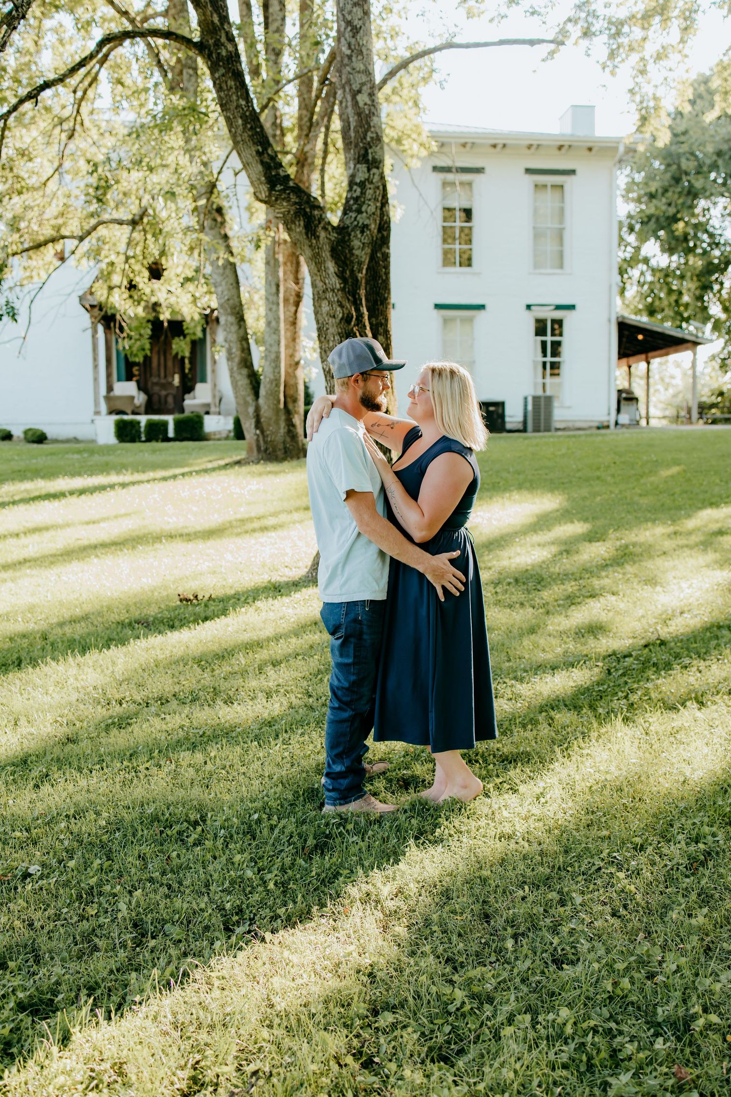 Couple embracing on green lawn in front of white historic, woman in navy dress and man in cap and jeans.