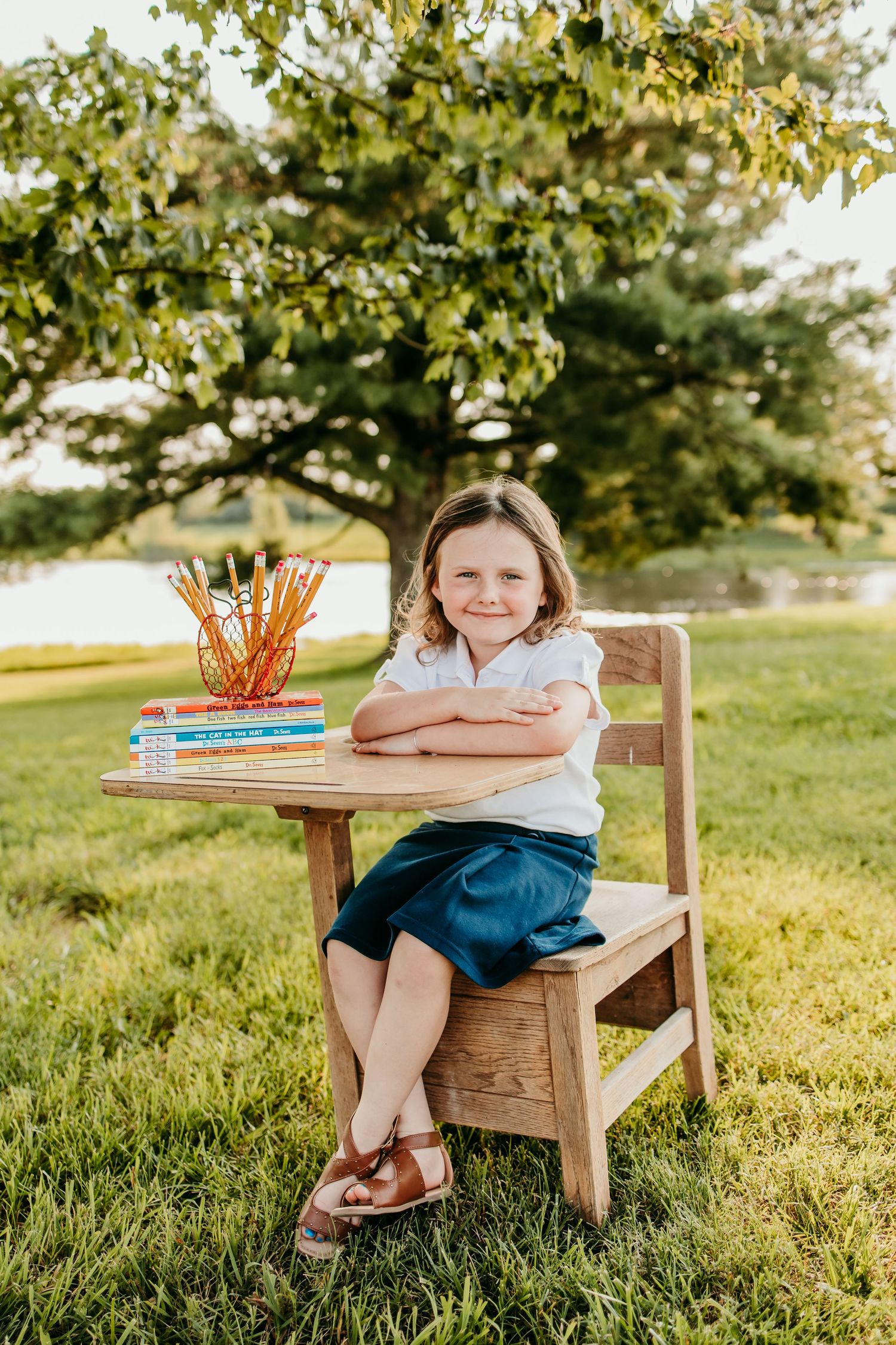 Young girl smiling at wooden school desk outdoors, with books and pencils, wearing white shirt and blue skirt.