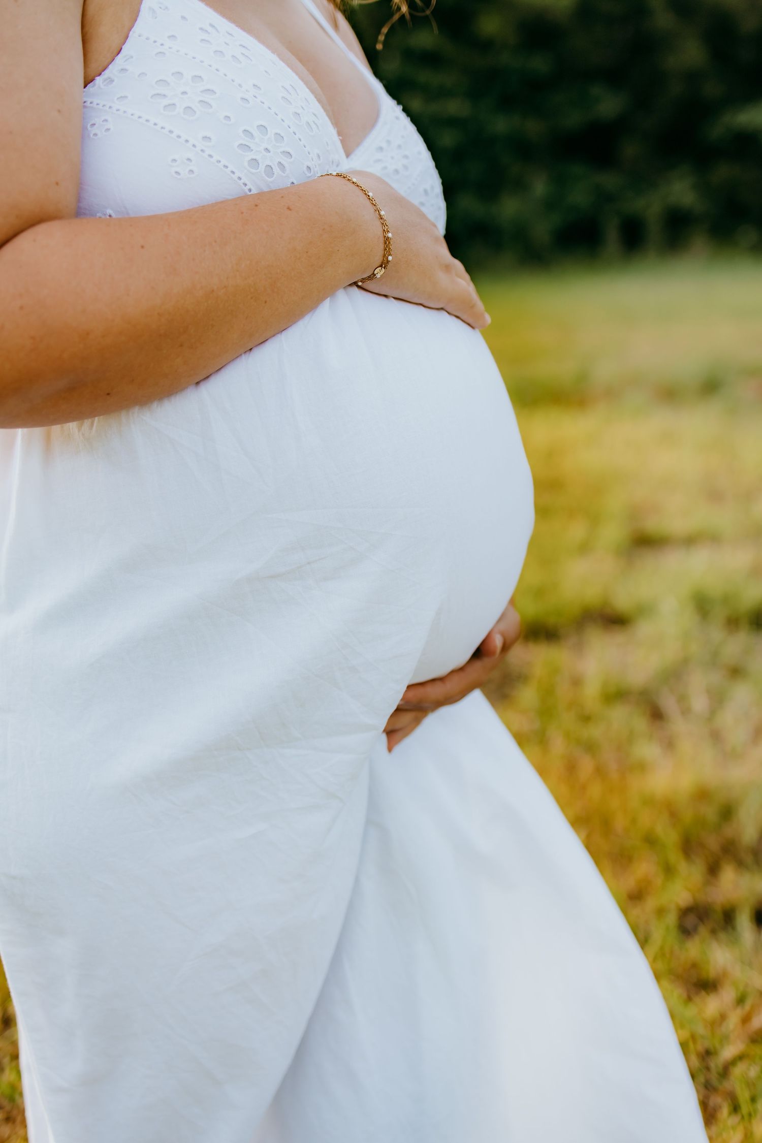 Maternity session in white dress cradling baby bump outdoors in a sunny field, wearing gold bracelet.