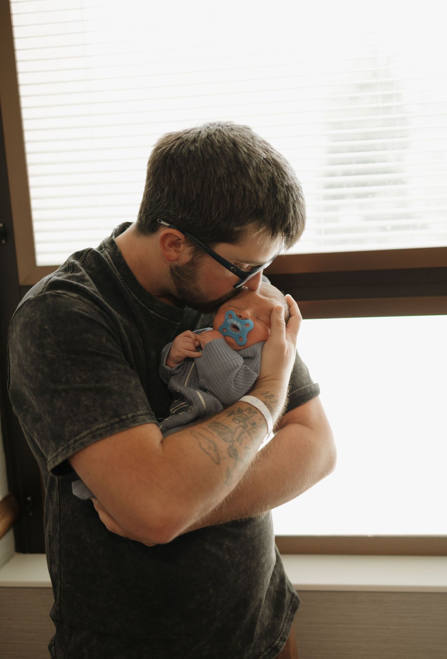 A tattooed father with glasses tenderly kisses his newborn baby near a bright hospital window.