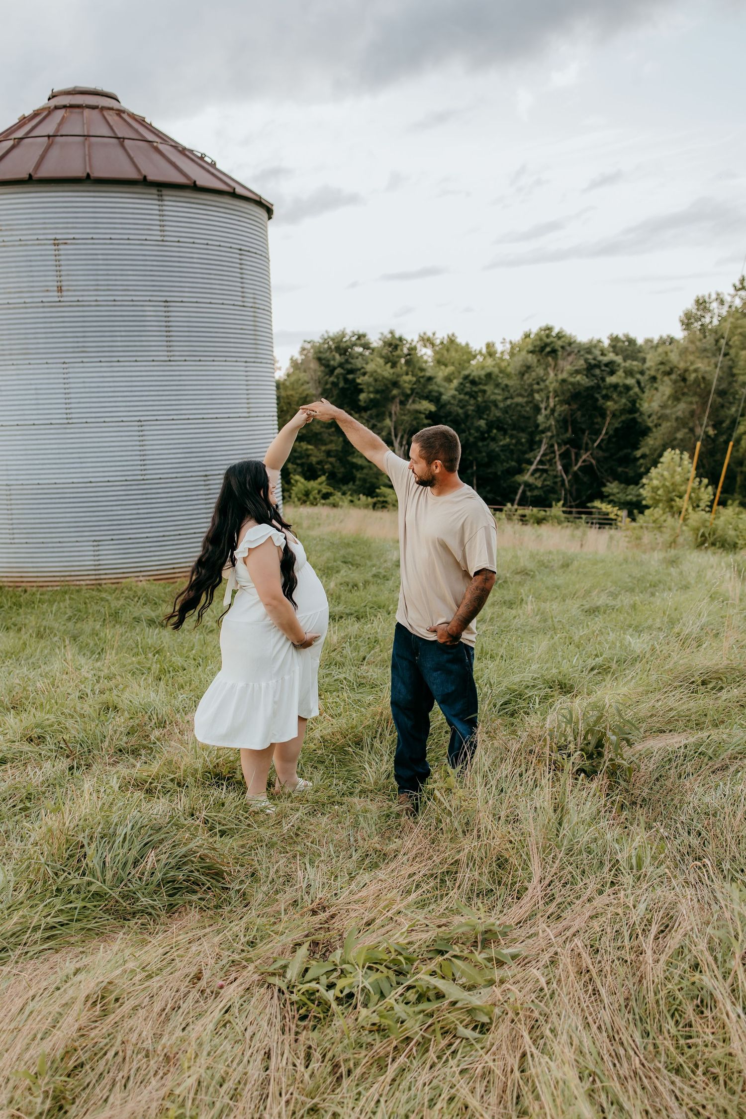 A couple dances in a grassy field near a grain silo, the woman in a white dress showing a baby bump.