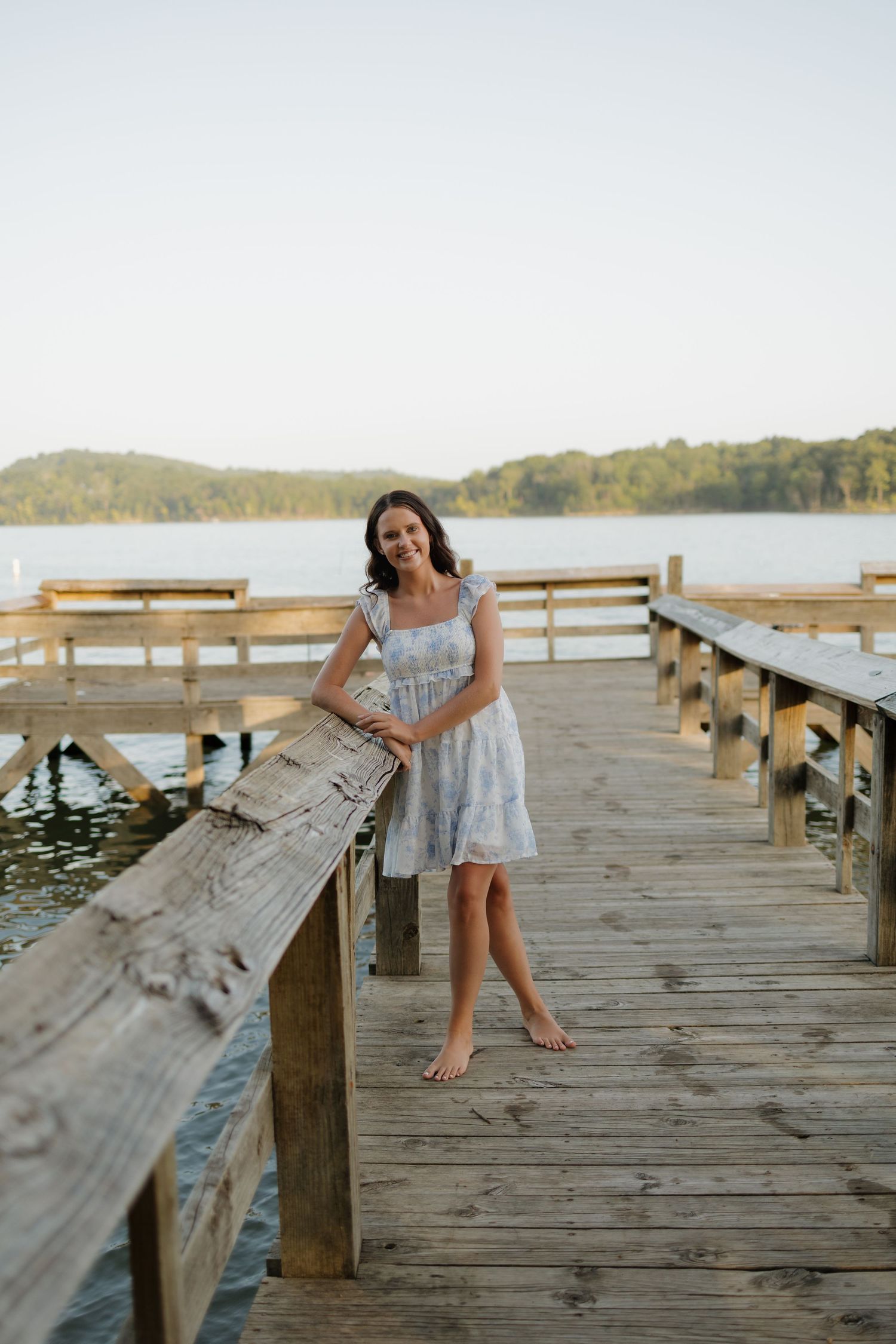 A barefoot woman in a light blue dress stands on a wooden dock by a calm lake surrounded by trees.