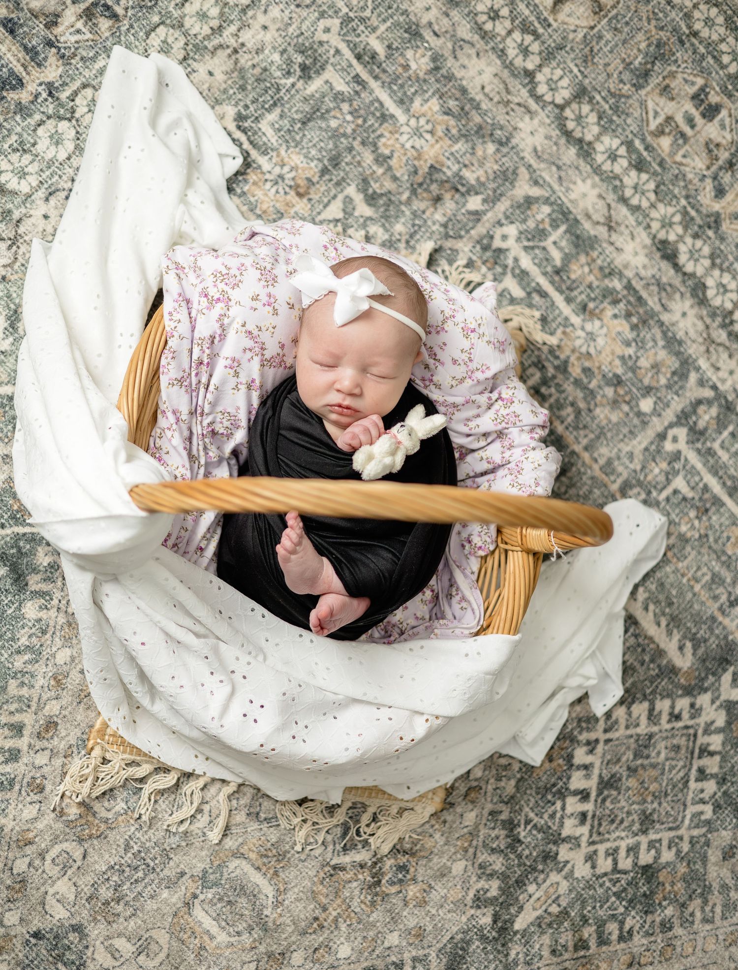 Newborn baby sleeping in a wicker basket wrapped in black swaddle with floral blanket and white bow headband.