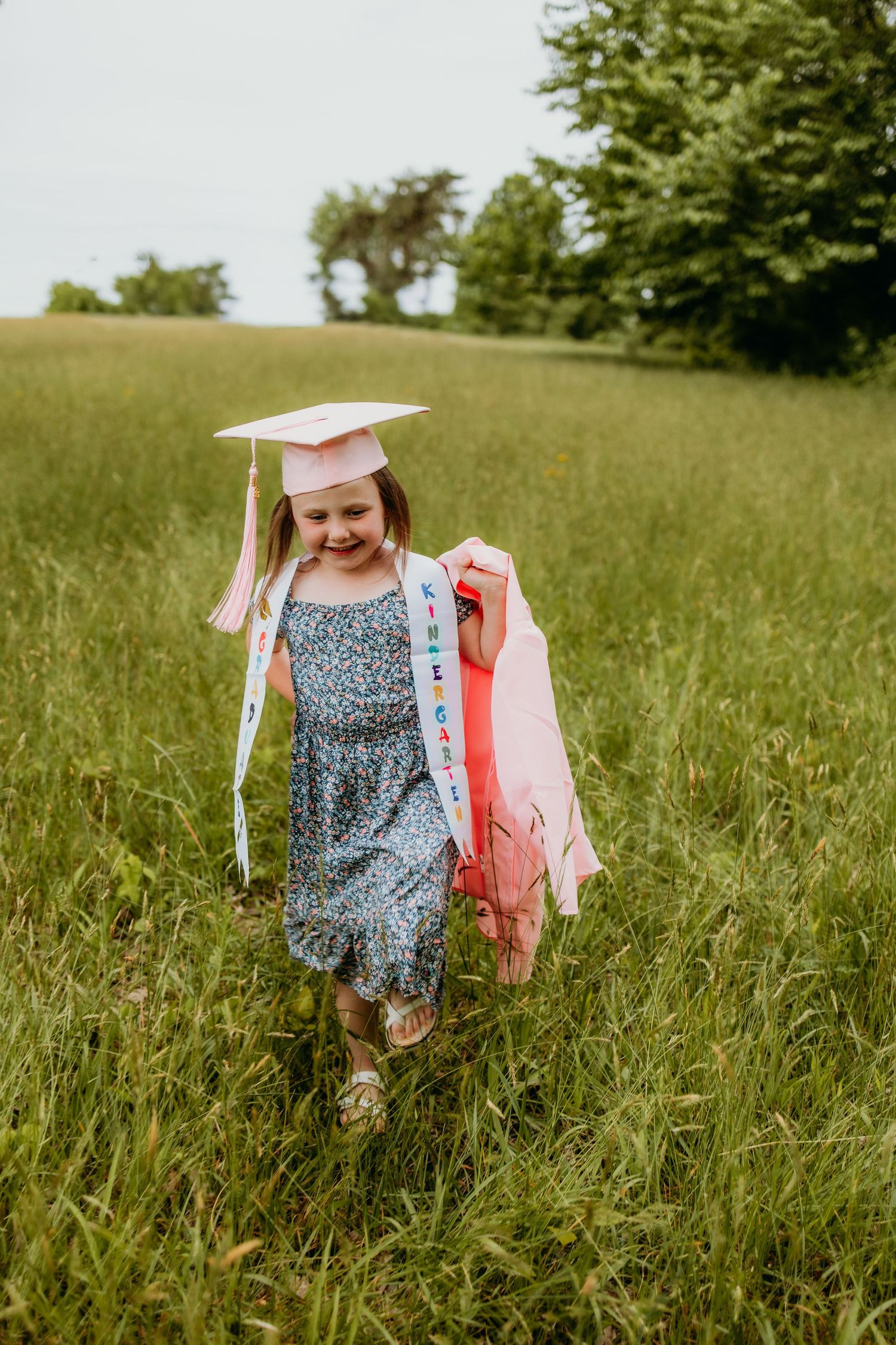 A young girl in a floral dress wearing a pink graduation cap and sash walks through a green meadow.