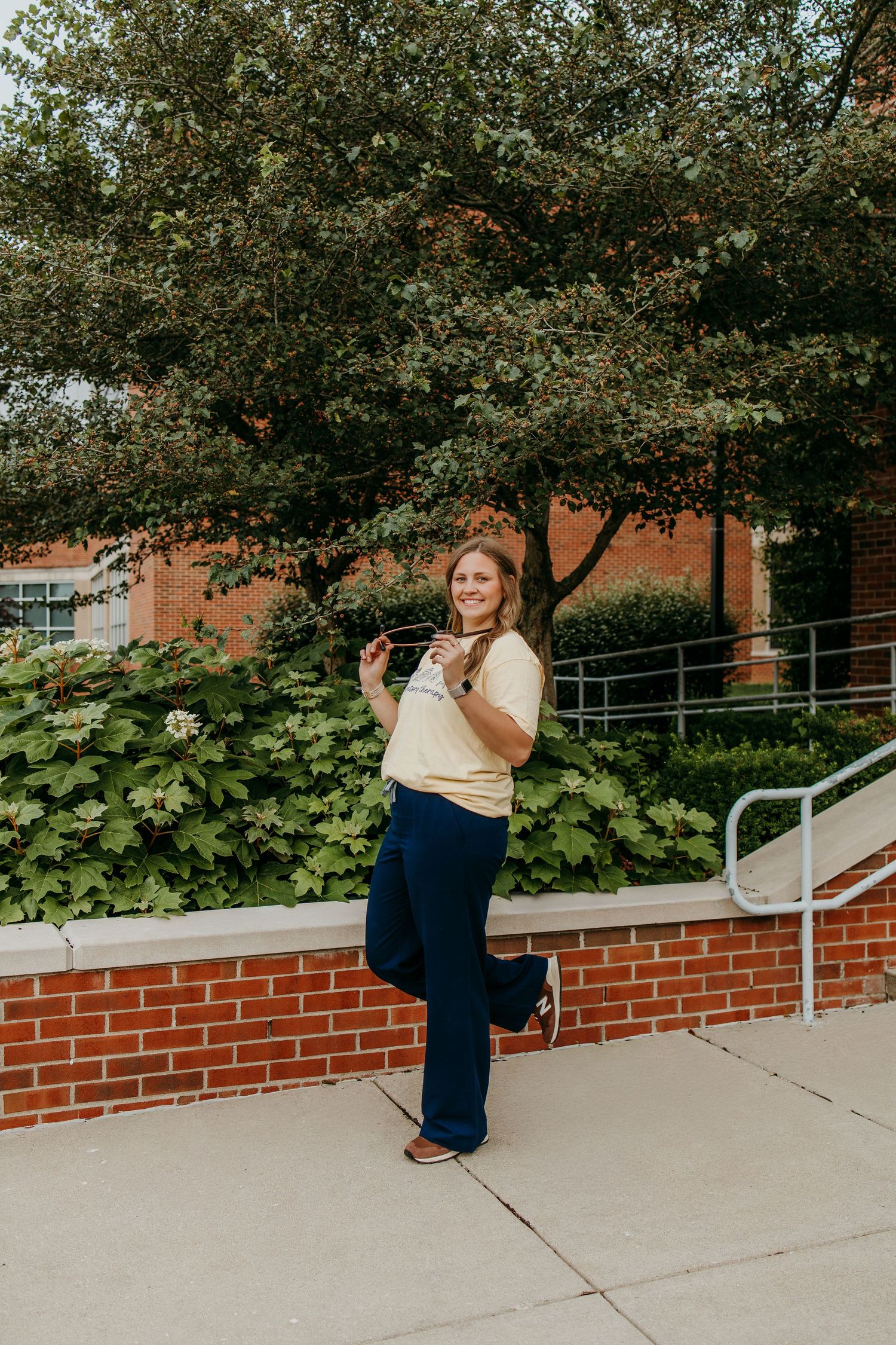 Young woman in yellow top and navy flare pants posing outdoors near brick wall with lush green trees behind her.