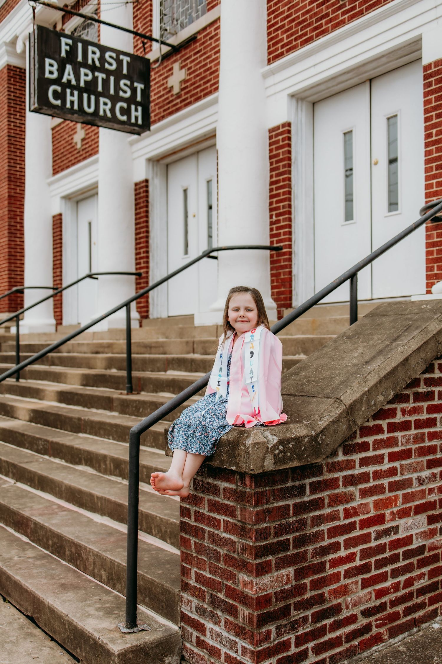 Young girl in pink cardigan sitting on brick steps outside First Baptist Church, smiling at camera.