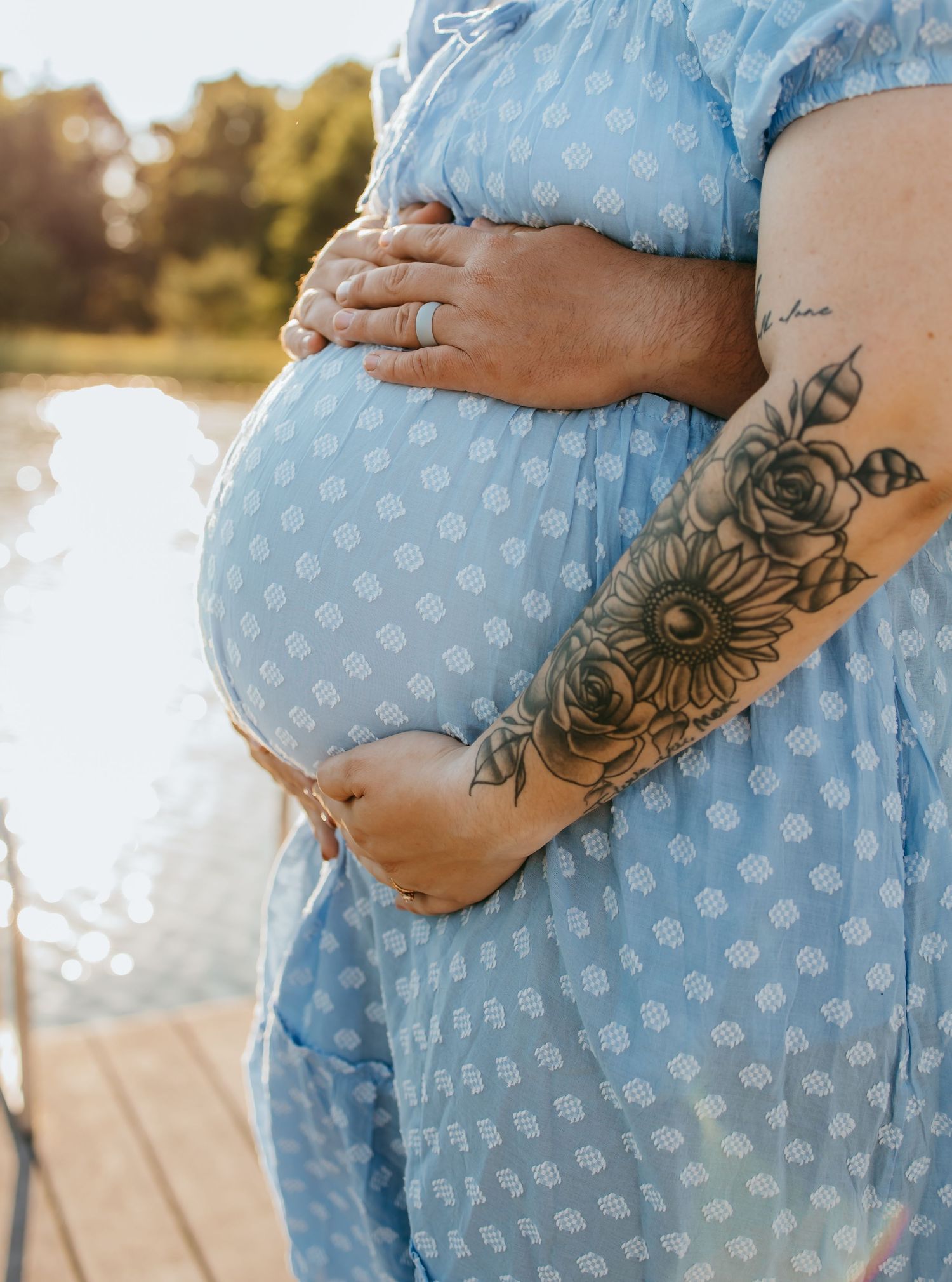 Pregnant woman in blue polka dot dress cradling baby bump, showcasing floral tattoo sleeve near a sunlit lake.