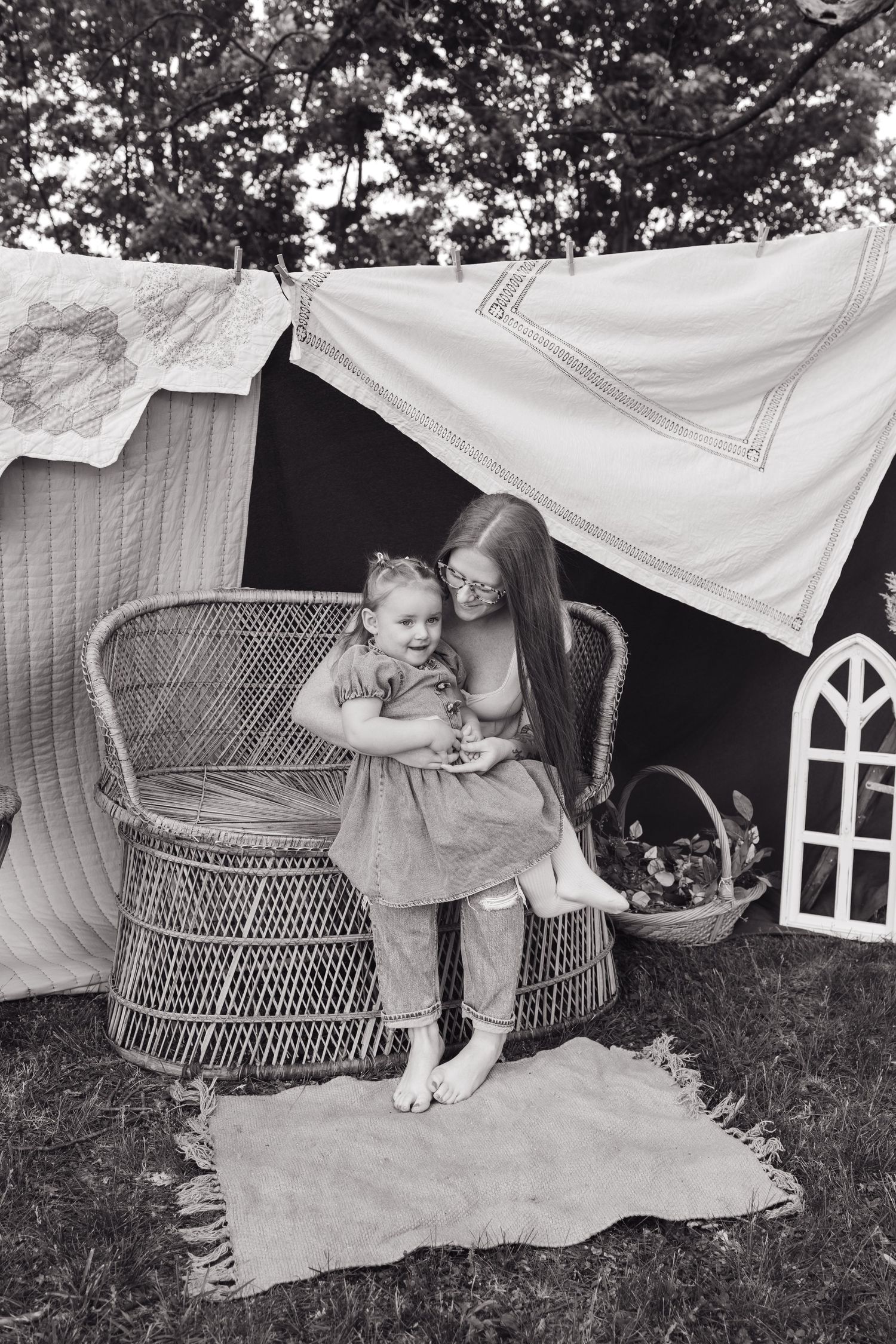 Black and white photo of a mother and toddler sitting in a wicker chair under a blanket fort outdoors.