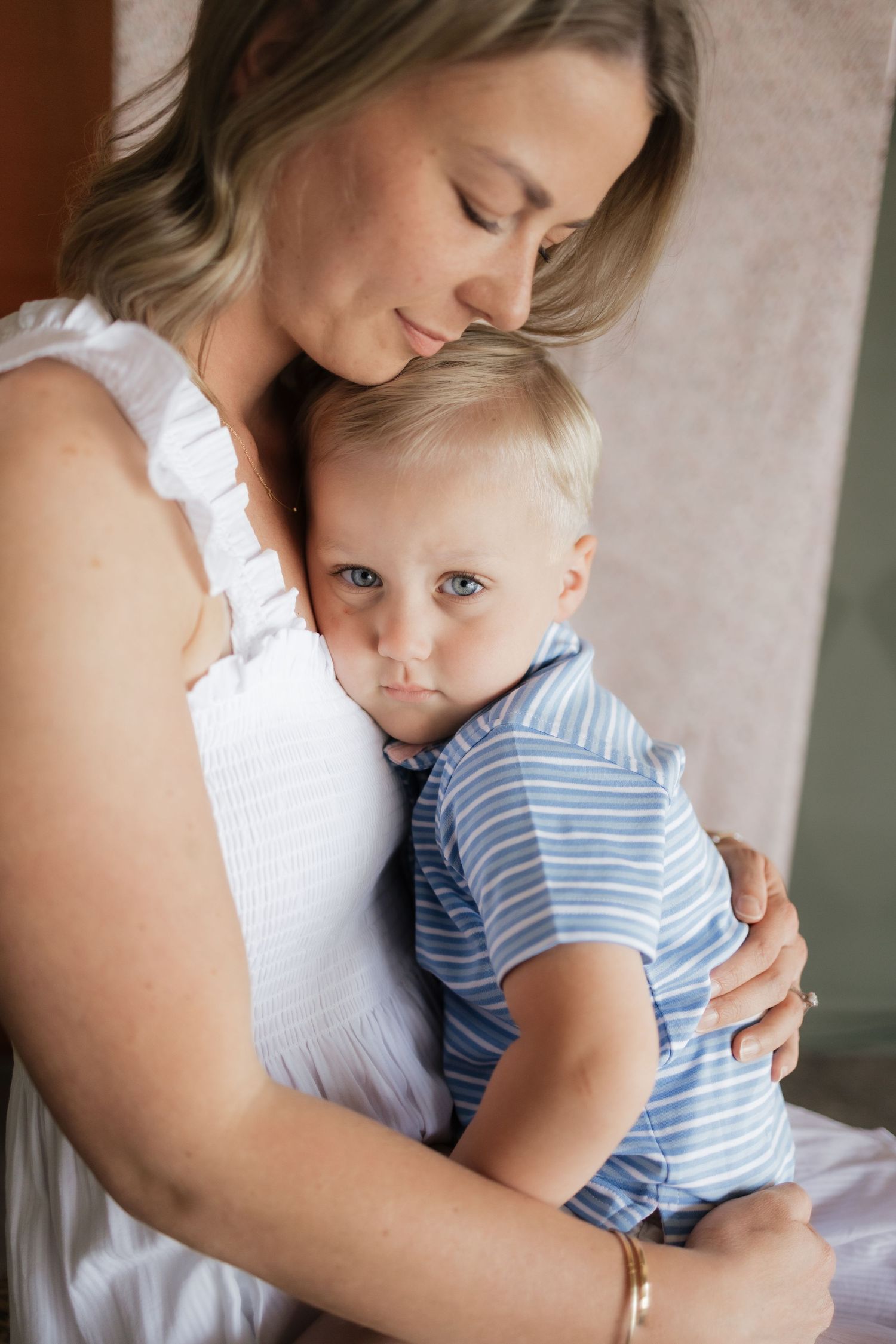 A smiling mother in a white dress tenderly embraces her young blond toddler in a blue striped outfit.
