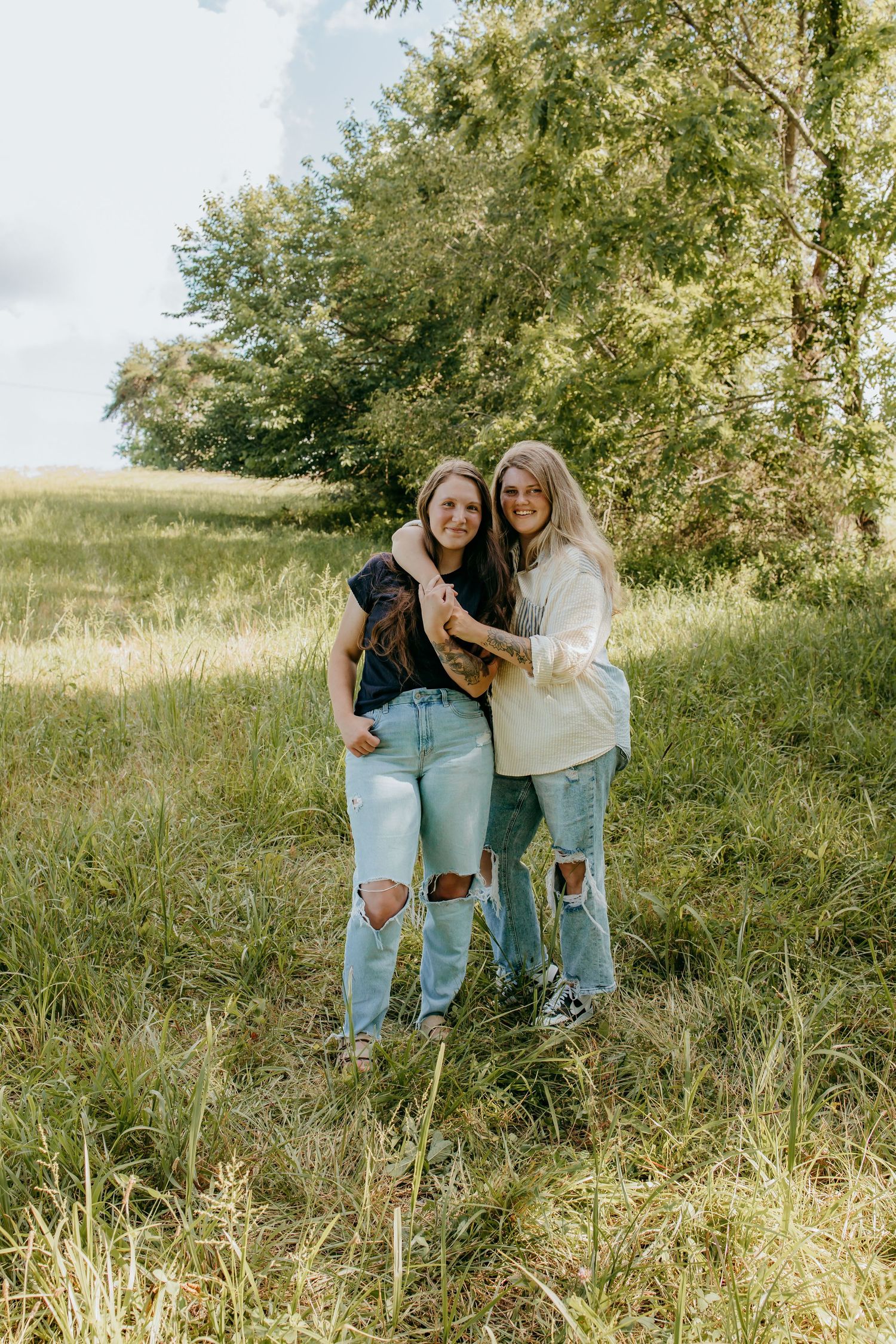 Two young women in ripped jeans embrace and smile in a lush green meadow with trees in the background.
