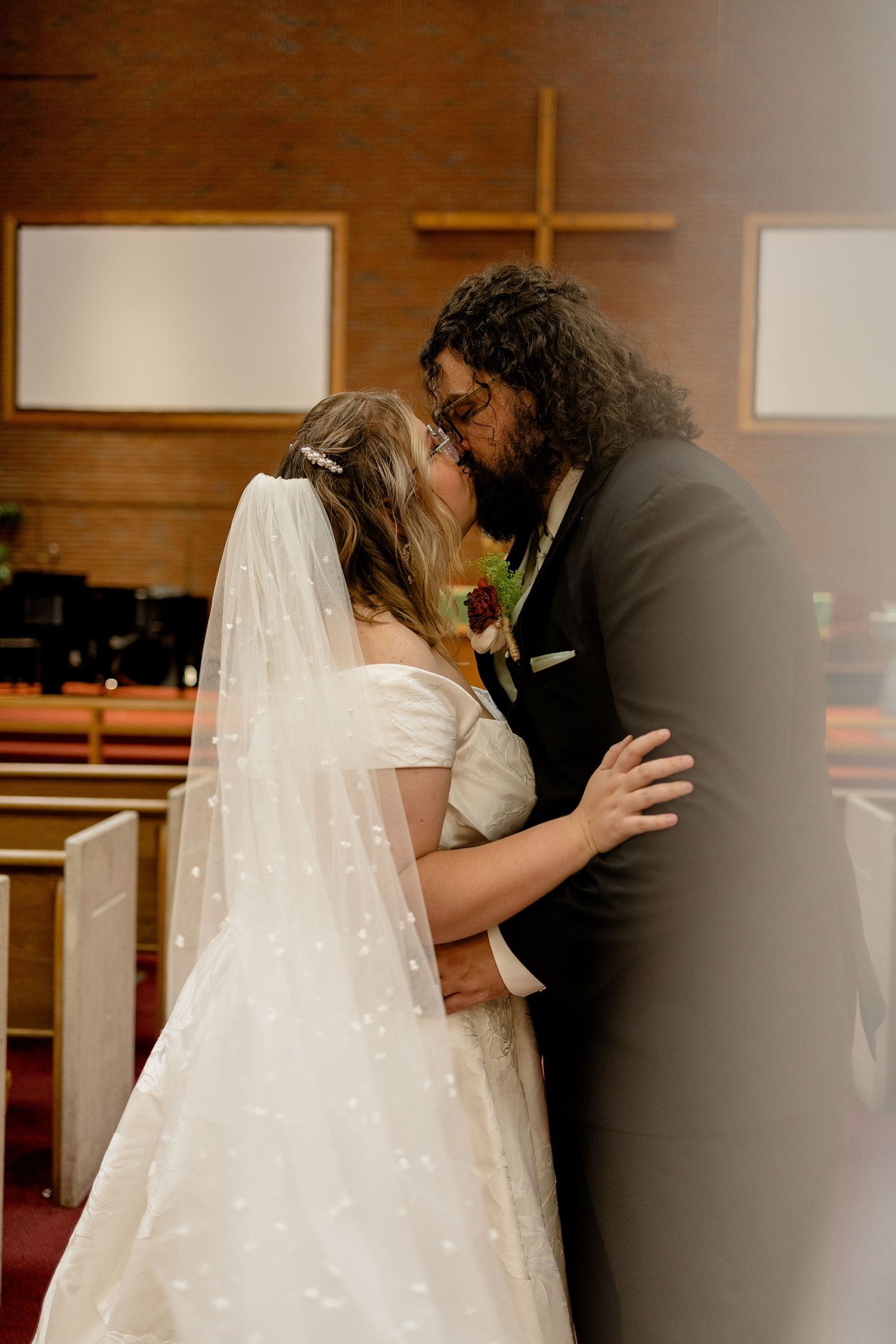 Bride and groom share first kiss at church altar, bride in white gown with veil, groom in dark suit with red boutonniere.
