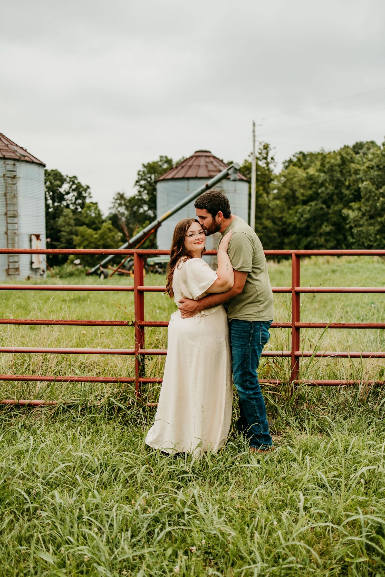 Couple embracing romantically in a lush green farm field with red gate and grain silos in background.