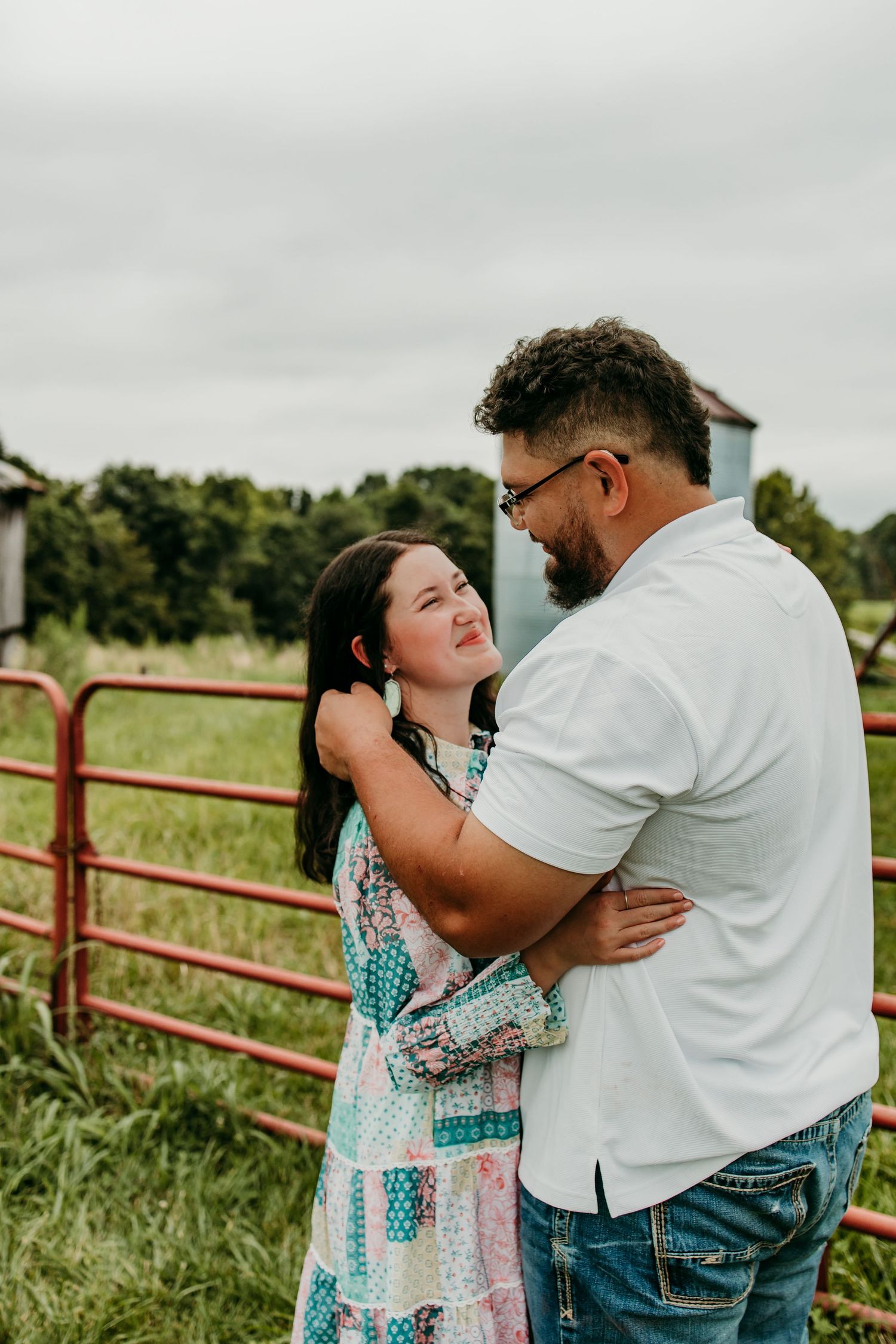 Couple embracing near red farm gate in green field, woman in patchwork dress smiling up at bearded man in white shirt.