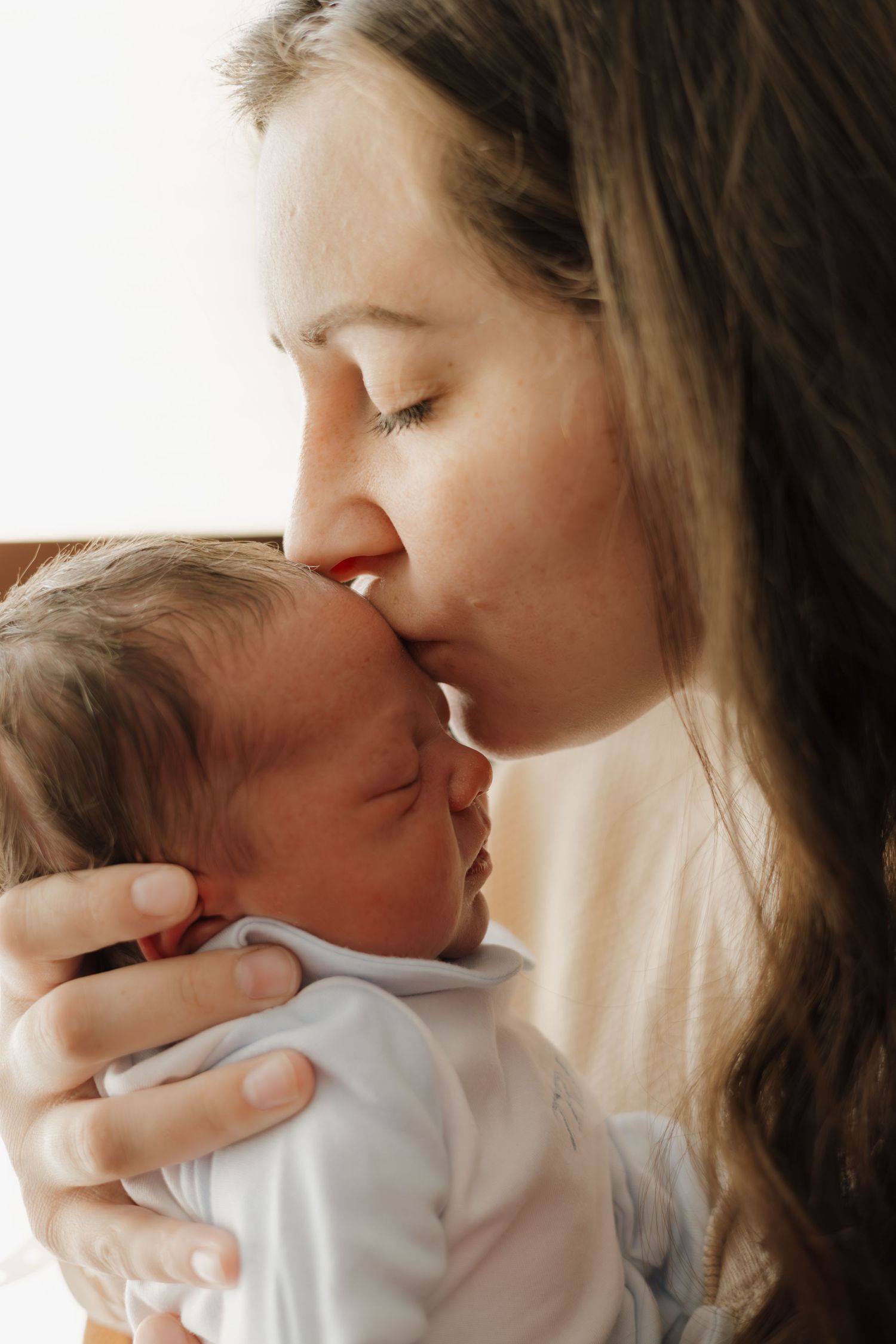 A tender moment as a young mother gently kisses her sleeping newborn baby on the forehead.