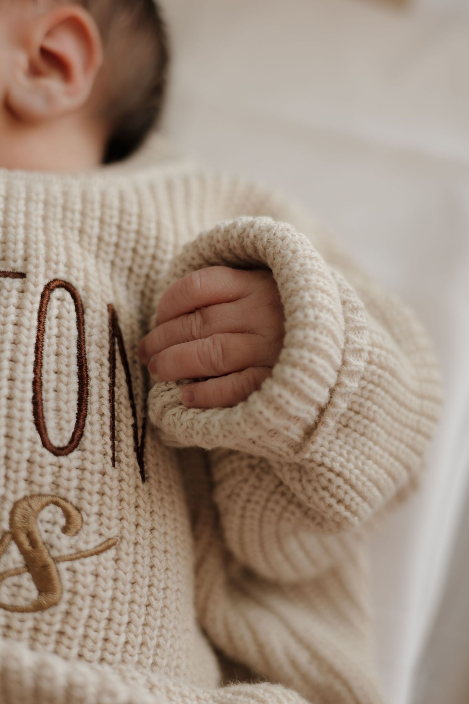 Close-up of a baby's tiny hand resting on a cream chunky knit sweater with embroidered lettering.