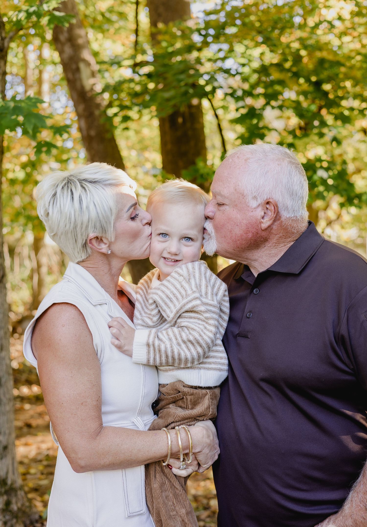 Grandparents kissing a smiling toddler on the cheeks outdoors in an autumn forest setting.