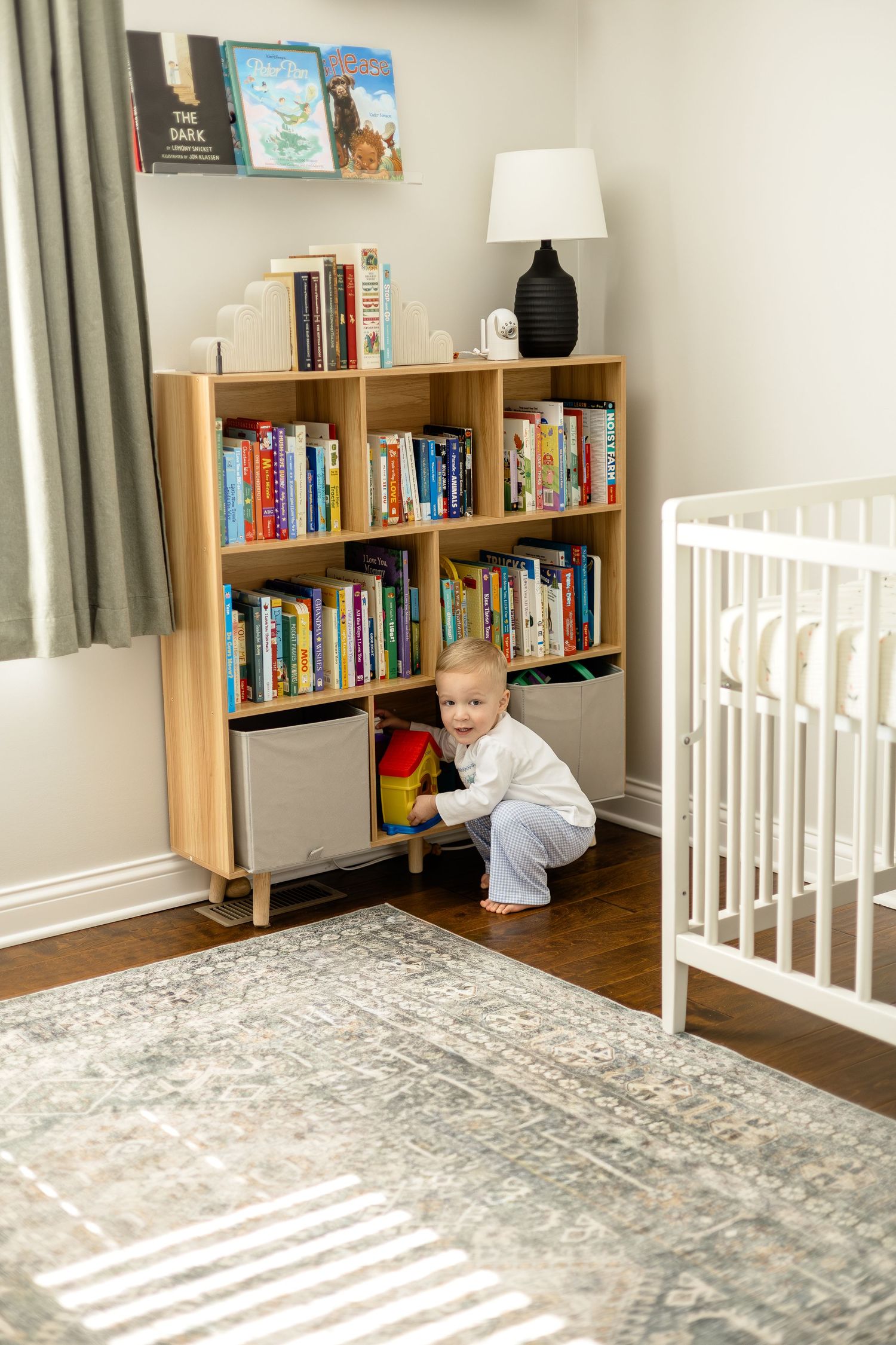Toddler crouching by a wooden bookshelf filled with children's books in a nursery with a white crib.