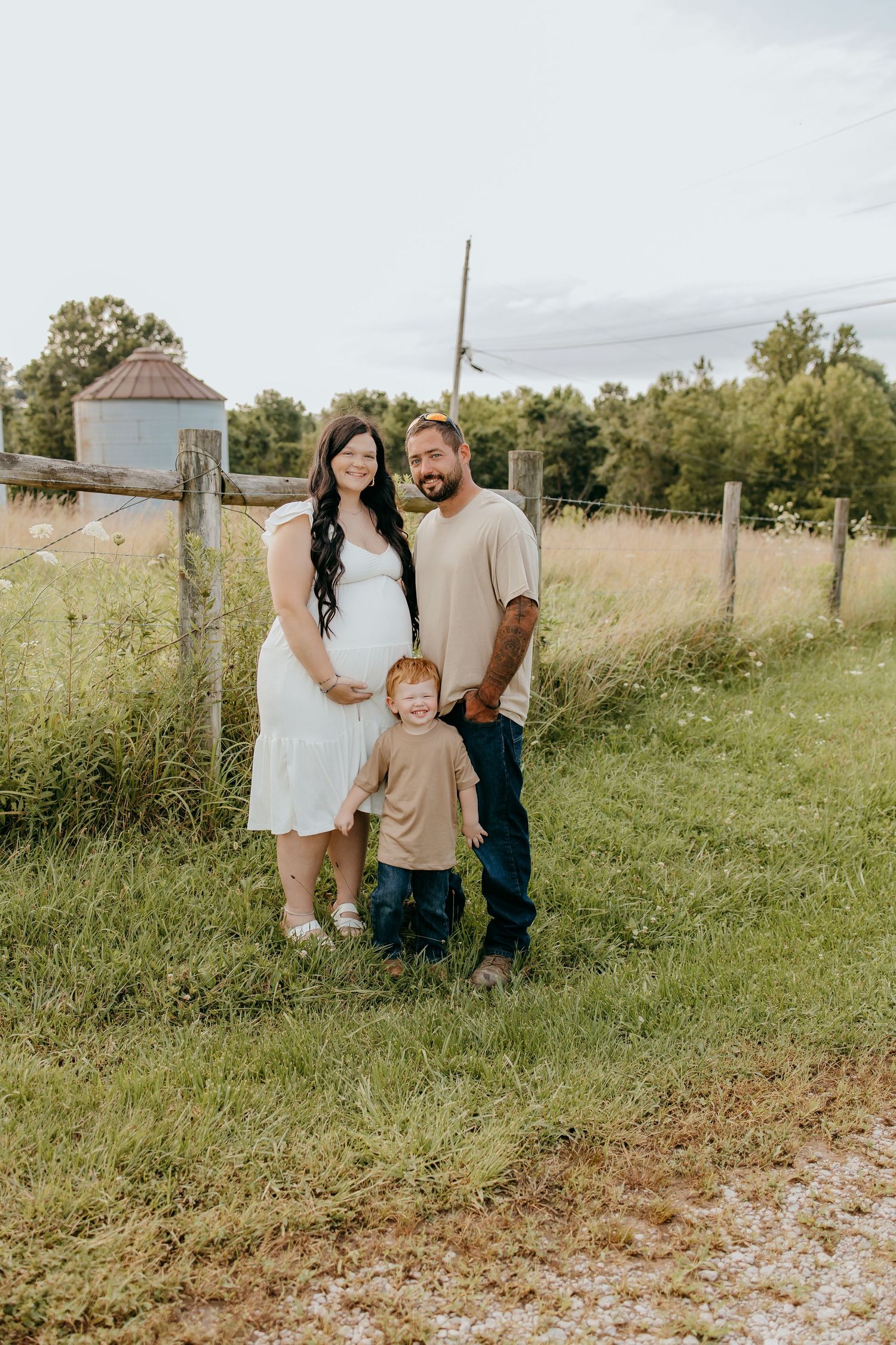 Pregnant woman in white dress with husband and toddler son posing on a rural farm path with green fields.