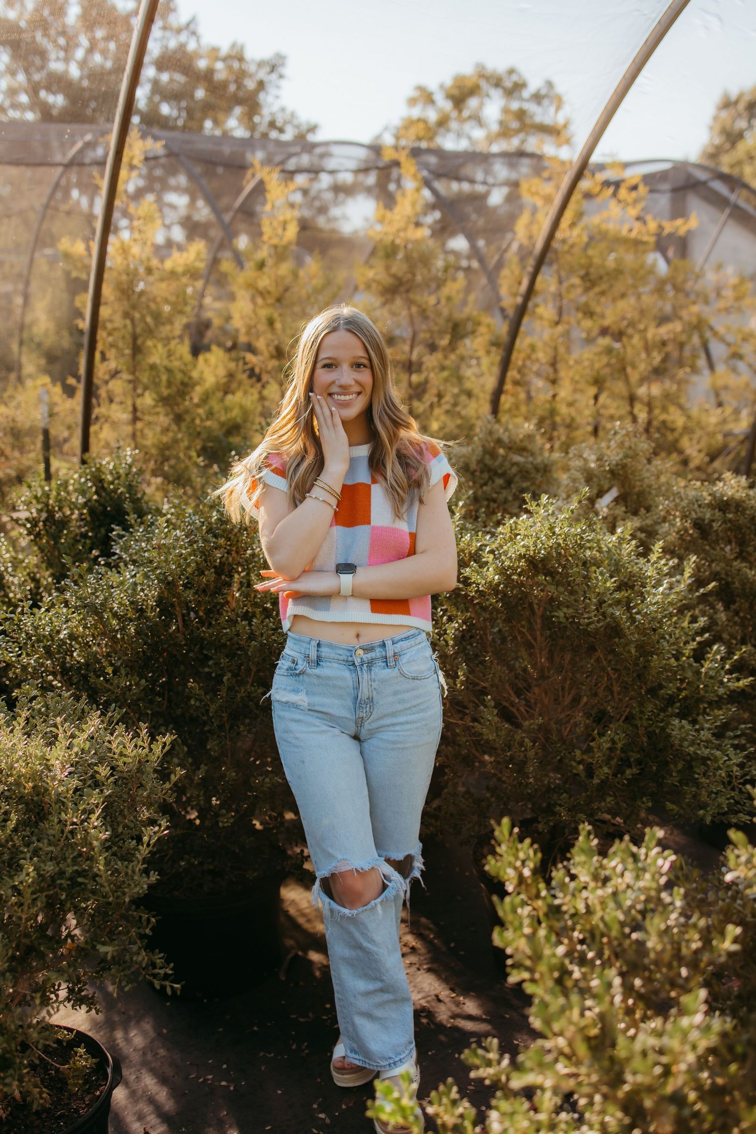 Smiling senior in colorful striped crop top and ripped jeans posing among green shrubs in sunny garden.