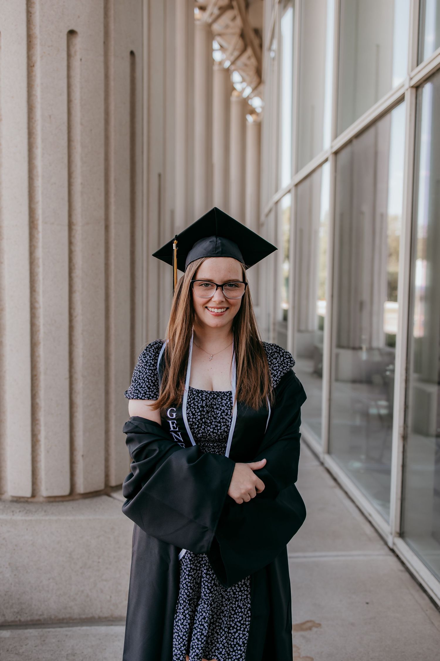 Smiling graduate in black cap and gown poses outdoors near stone columns, wearing glasses and floral dress.